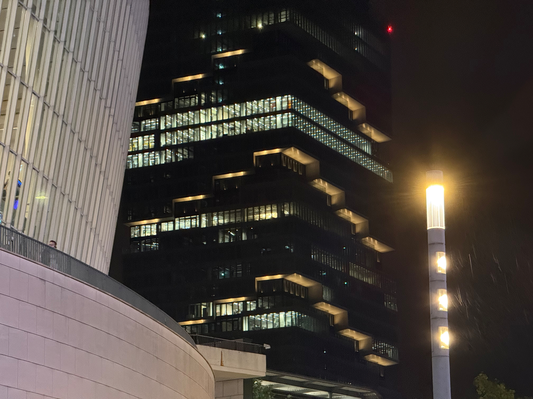 A modern, multi-story building with illuminated windows and a streetlamp glowing brightly in the foreground during nighttime.