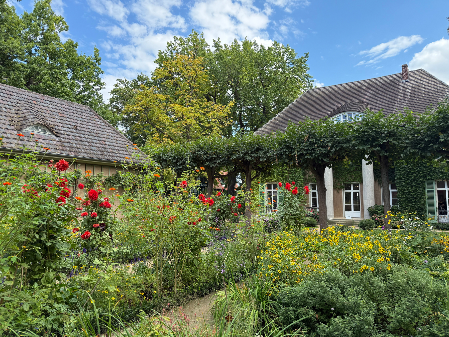 A charming garden full of vibrant flowers and greenery surrounds a traditional summer house and garden cottage with sloped roofs and an arched window.