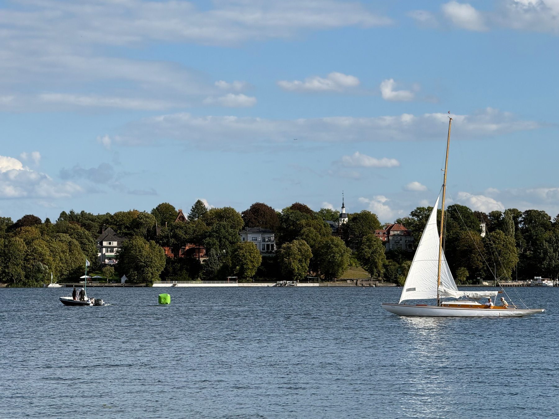 A sailboat and a motorboat are on a calm body of water near a shoreline lined with trees and houses under a partly cloudy sky.