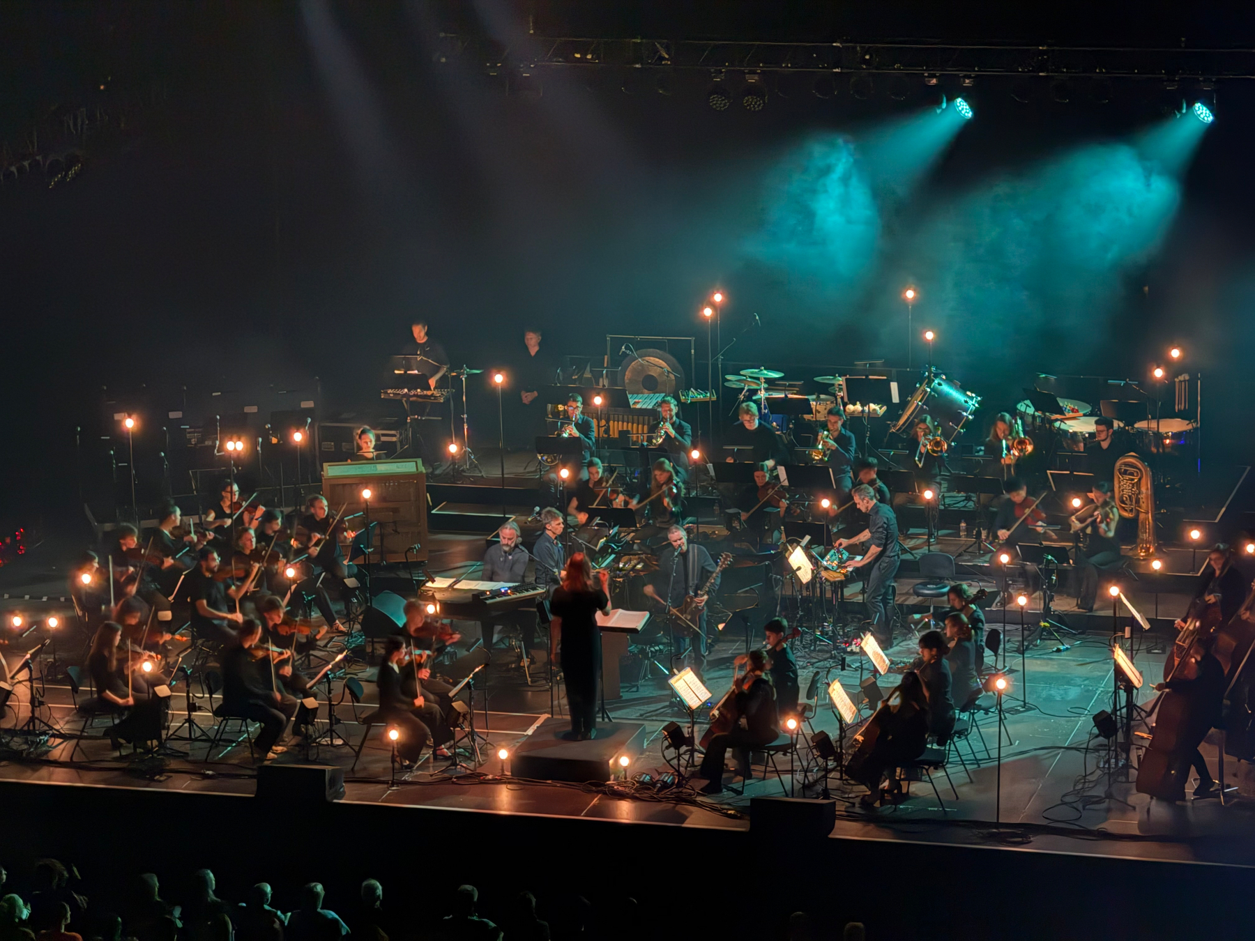 A conductor leads an orchestra performing on a dimly lit stage with musicians playing various instruments surrounded by small individual lights.