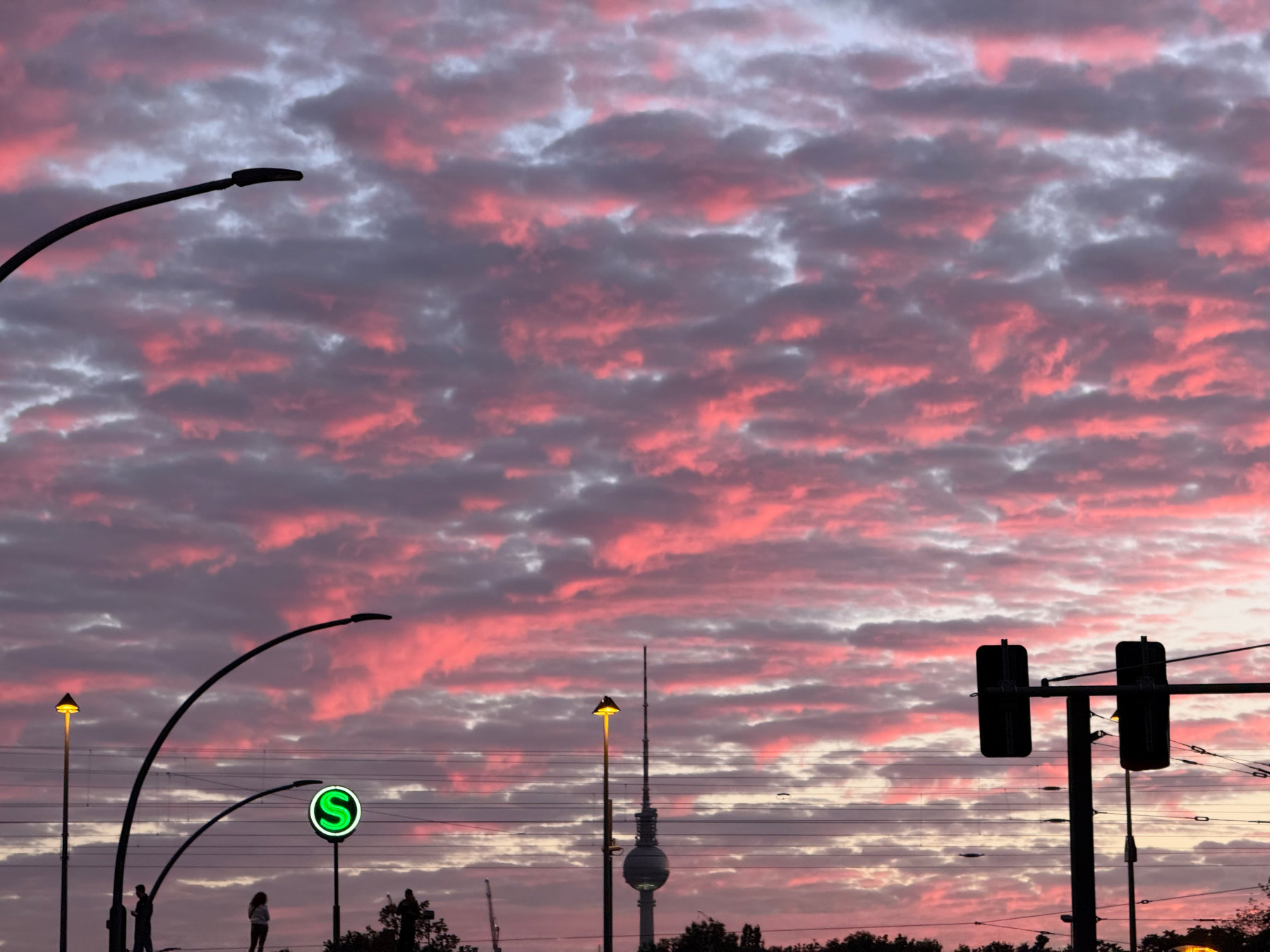 A vibrant pink and purple sunset sky serves as a backdrop to streetlights, traffic signals, and the tall Berlin Fernsehturm tv tower with a spherical observation deck.