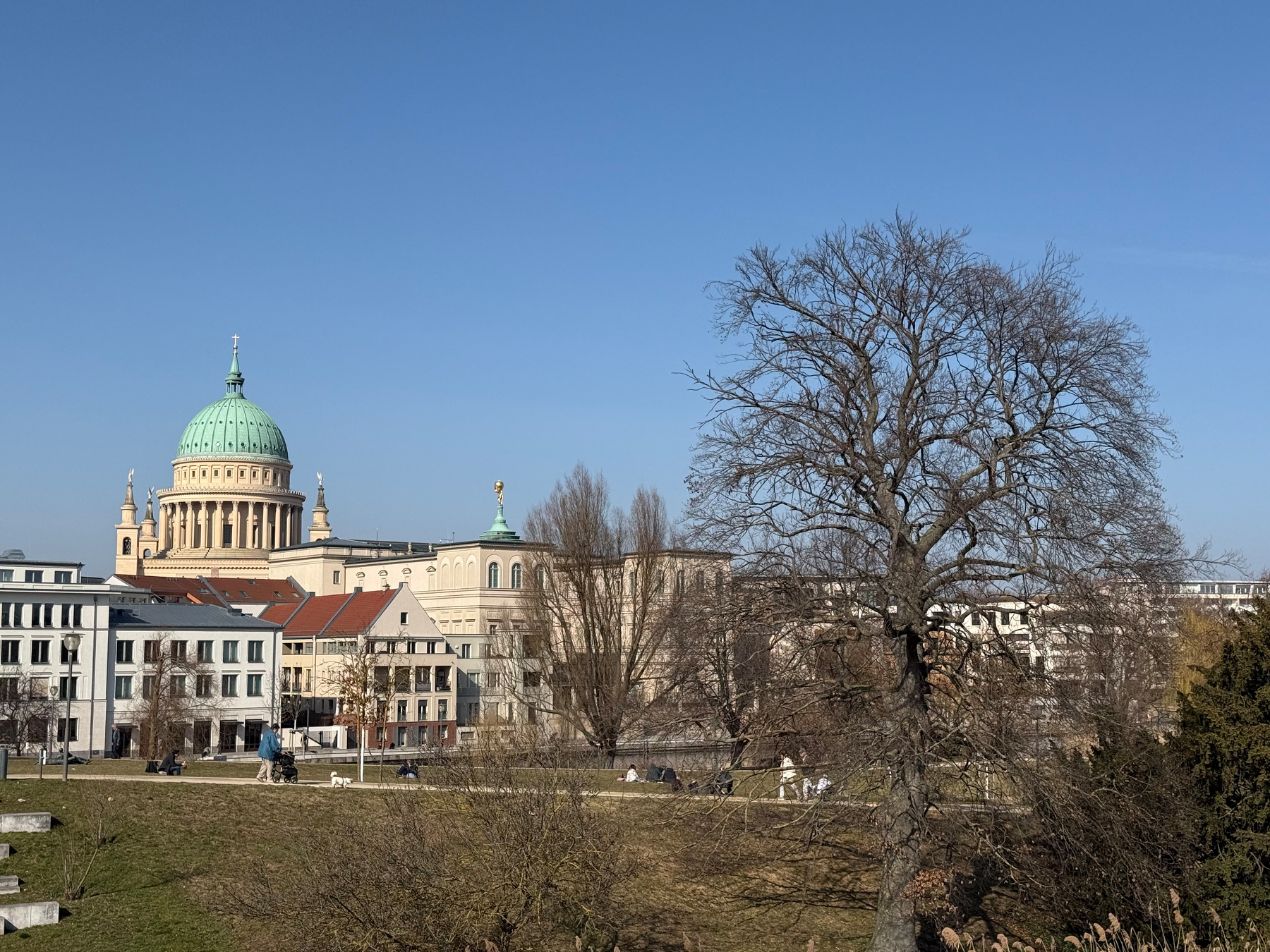 A historic building with a large green dome rises against a clear blue sky, surrounded by trees and smaller structures.