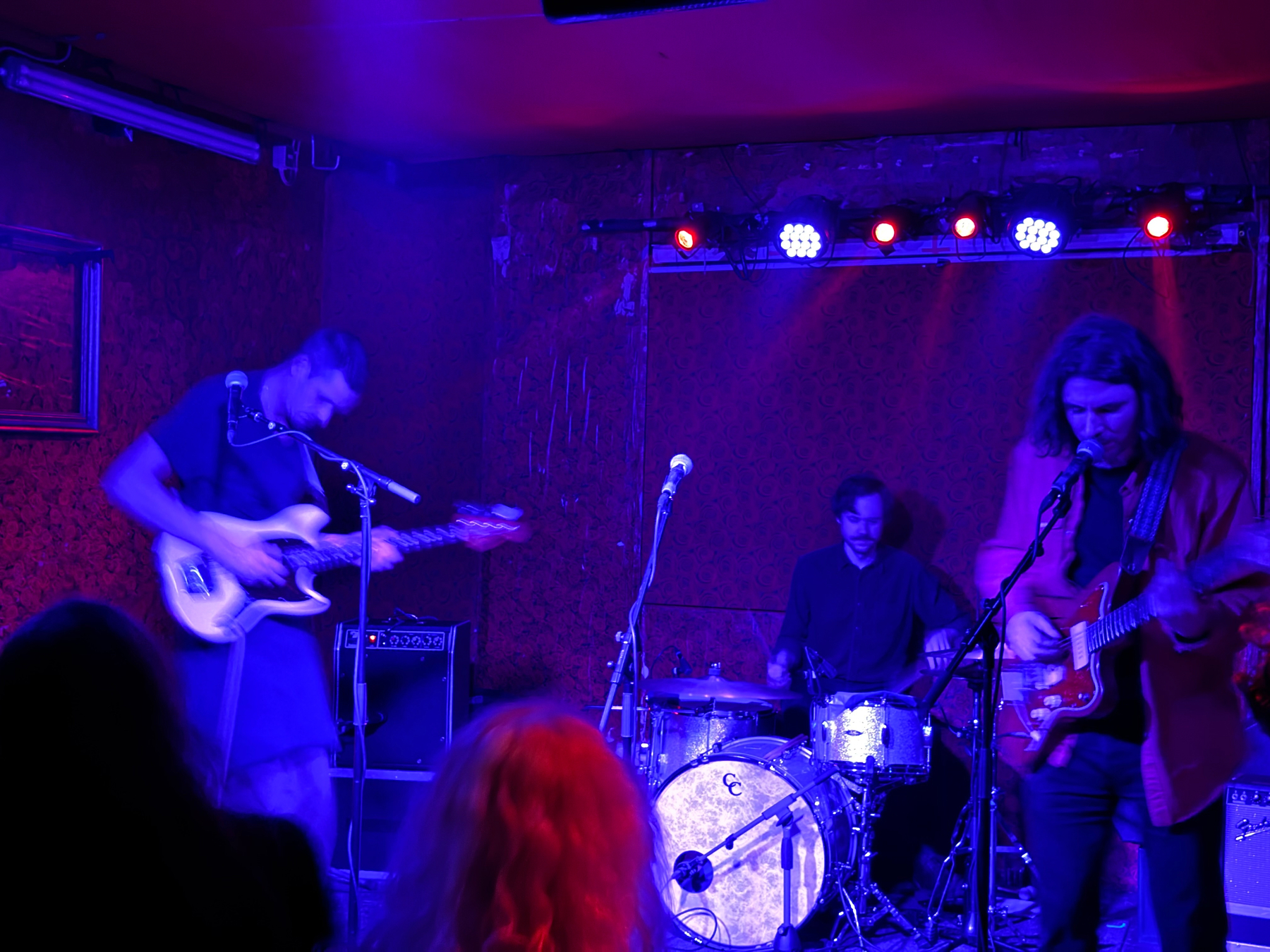 A band is performing on stage in a dimly lit venue, with blue and purple lighting. Bass player on the left, drummer in the middle, singer on electric guitar on the right