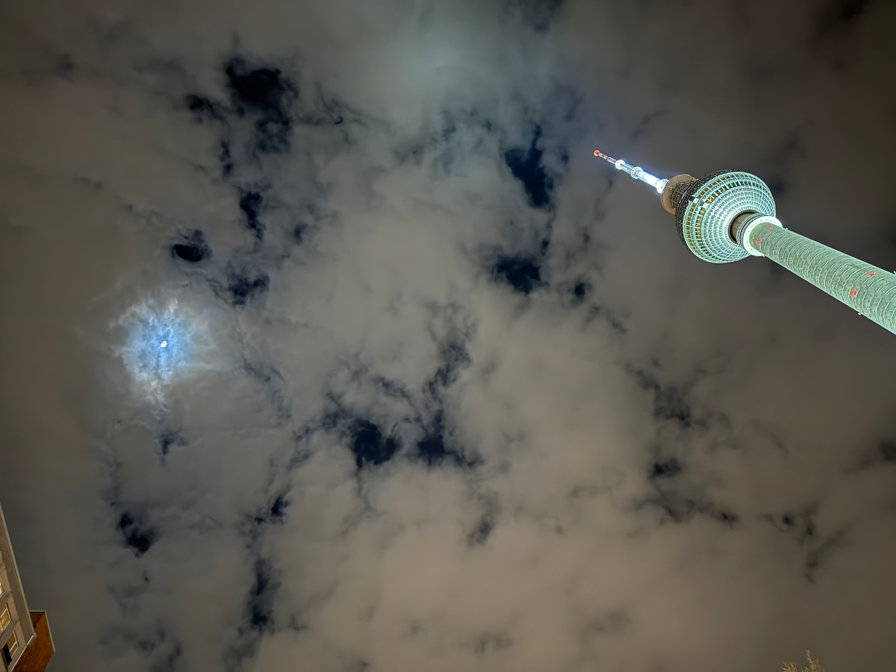 A tall, illuminated tower, Berlin Fernsehturm, extends right into the cloudy night sky with a bright moon partially visible behind the clouds.