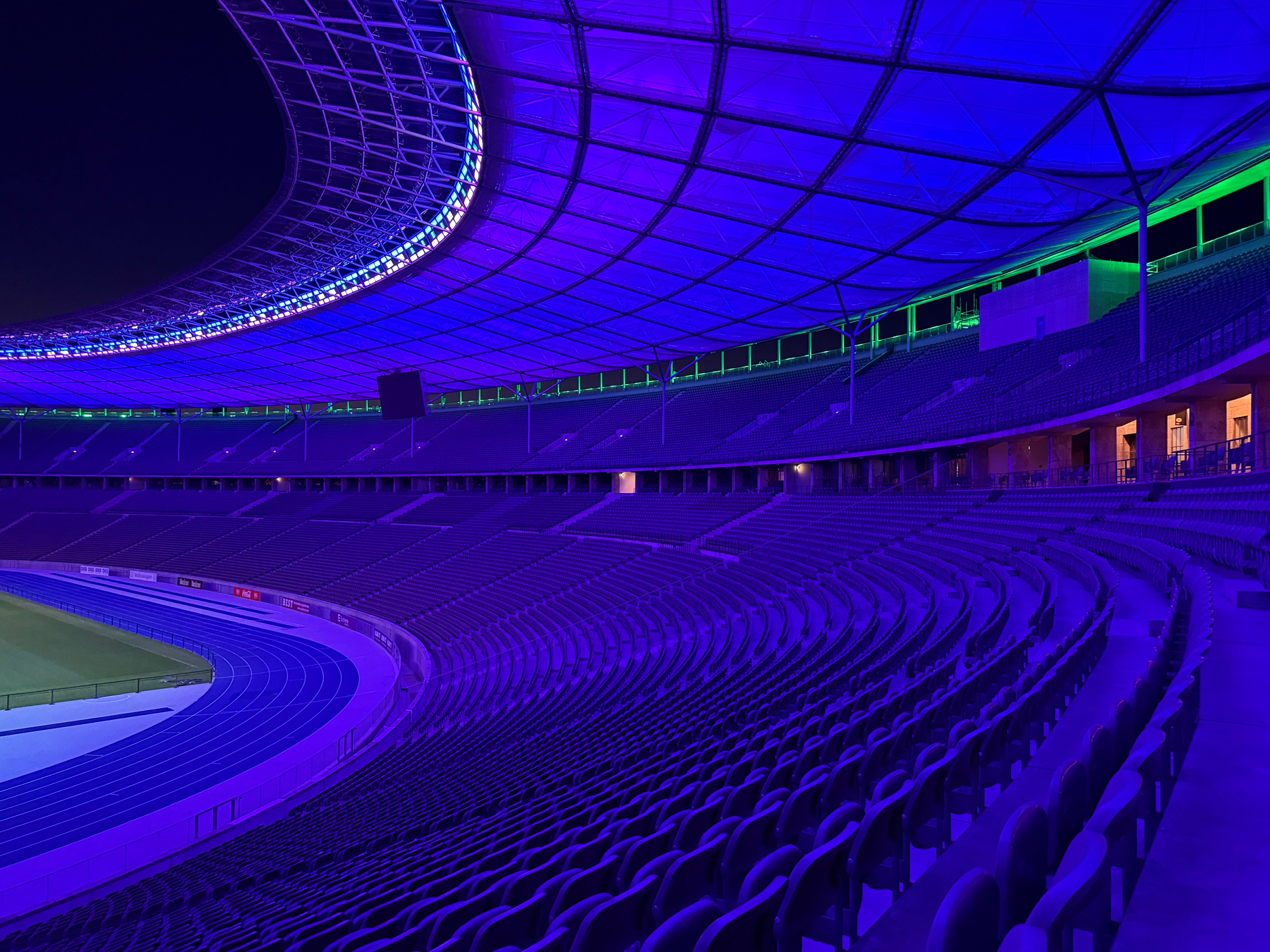 A stadium with blue and purple lighting illuminates rows of empty seating surrounding a running track and field.