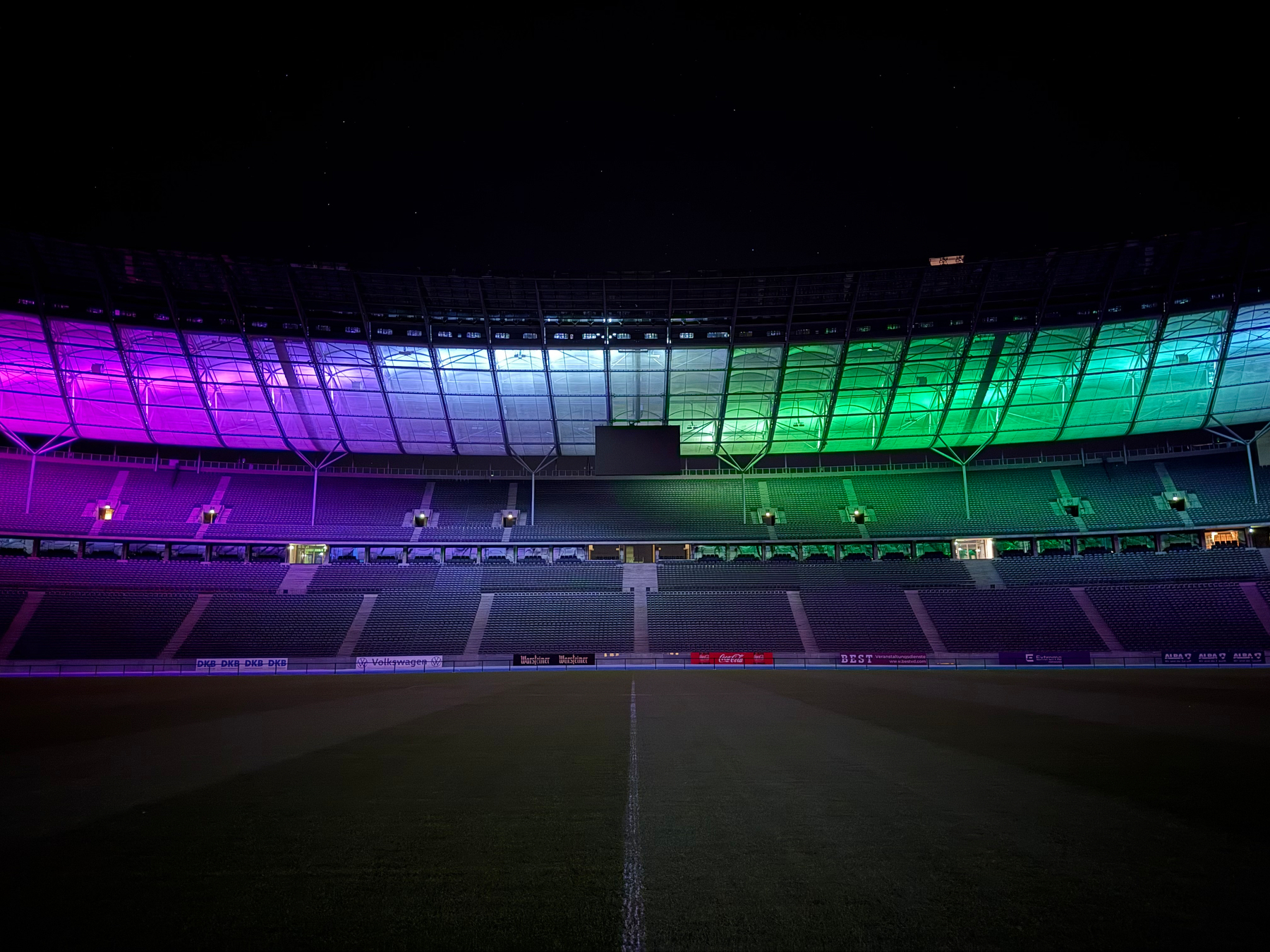 A stadium with empty field is lit at night with colorful lights in shades of purple, blue, and green illuminating the empty seating area.