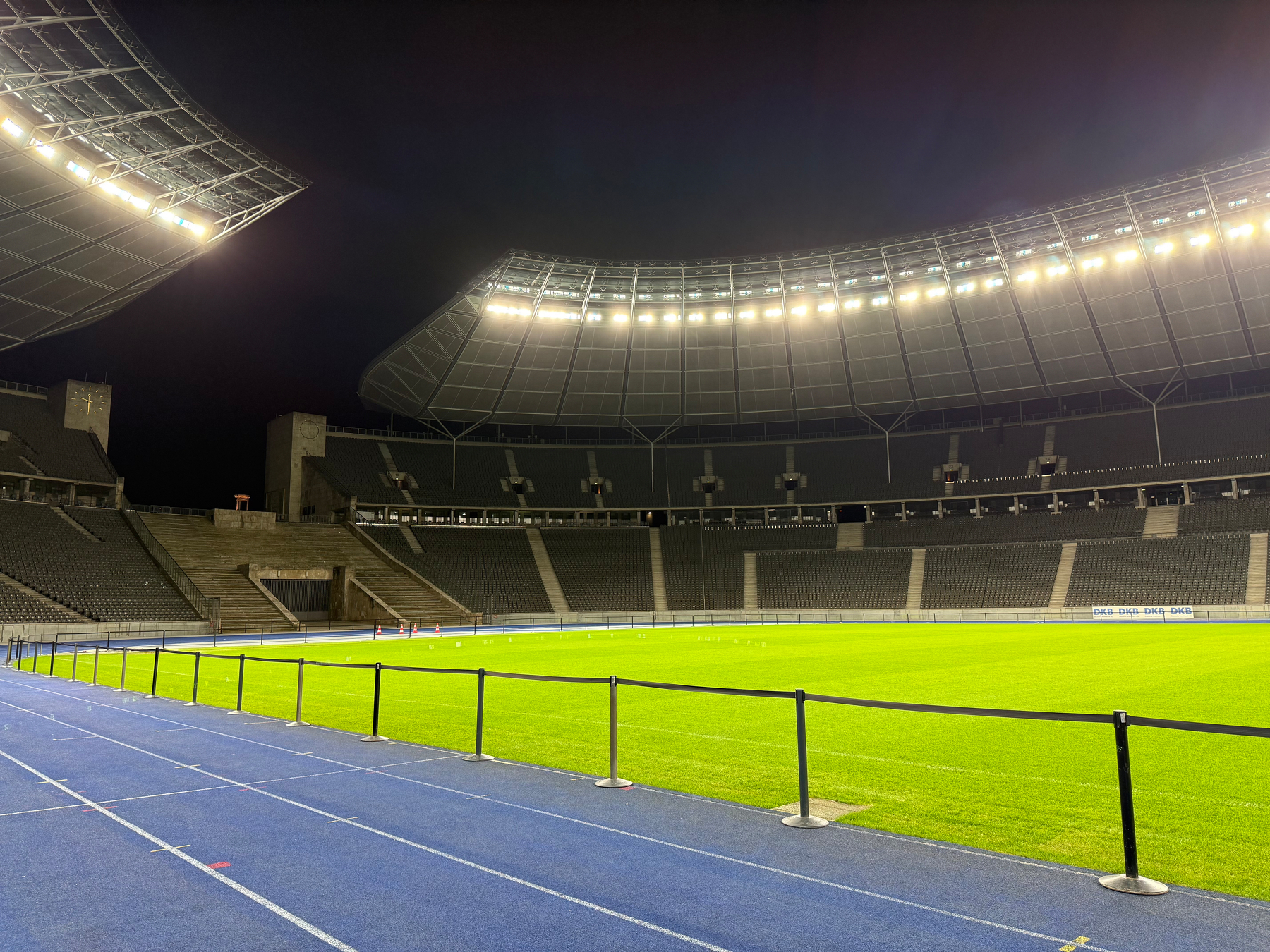 A brightly lit sports stadium with an expansive, empty field is surrounded by rows of empty seats and a blue running track.