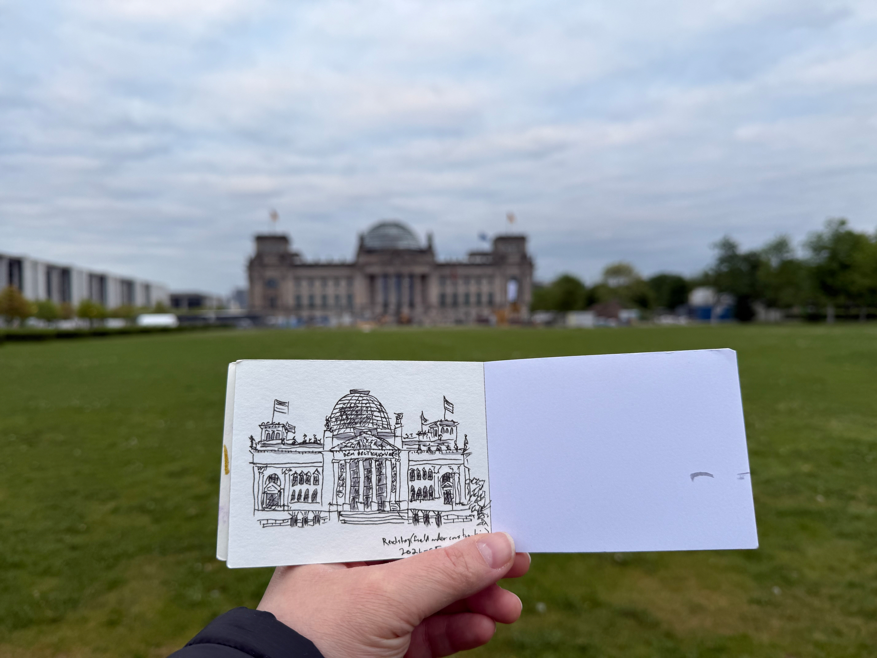 A hand  holds a sketchbook featuring a drawing of the Reichstag dome-topped building, matching the real-life building in the background.