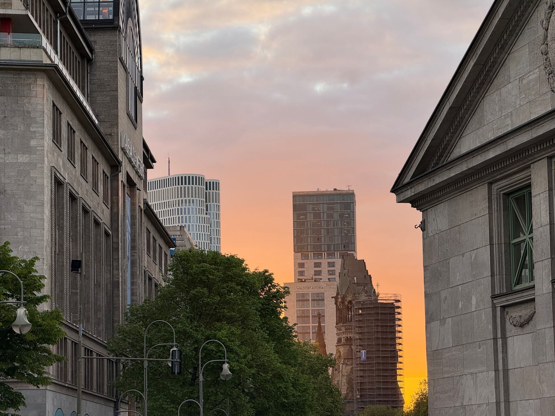 A cityscape features tall buildings against a vibrant evening sky with trees and street lights in the foreground.