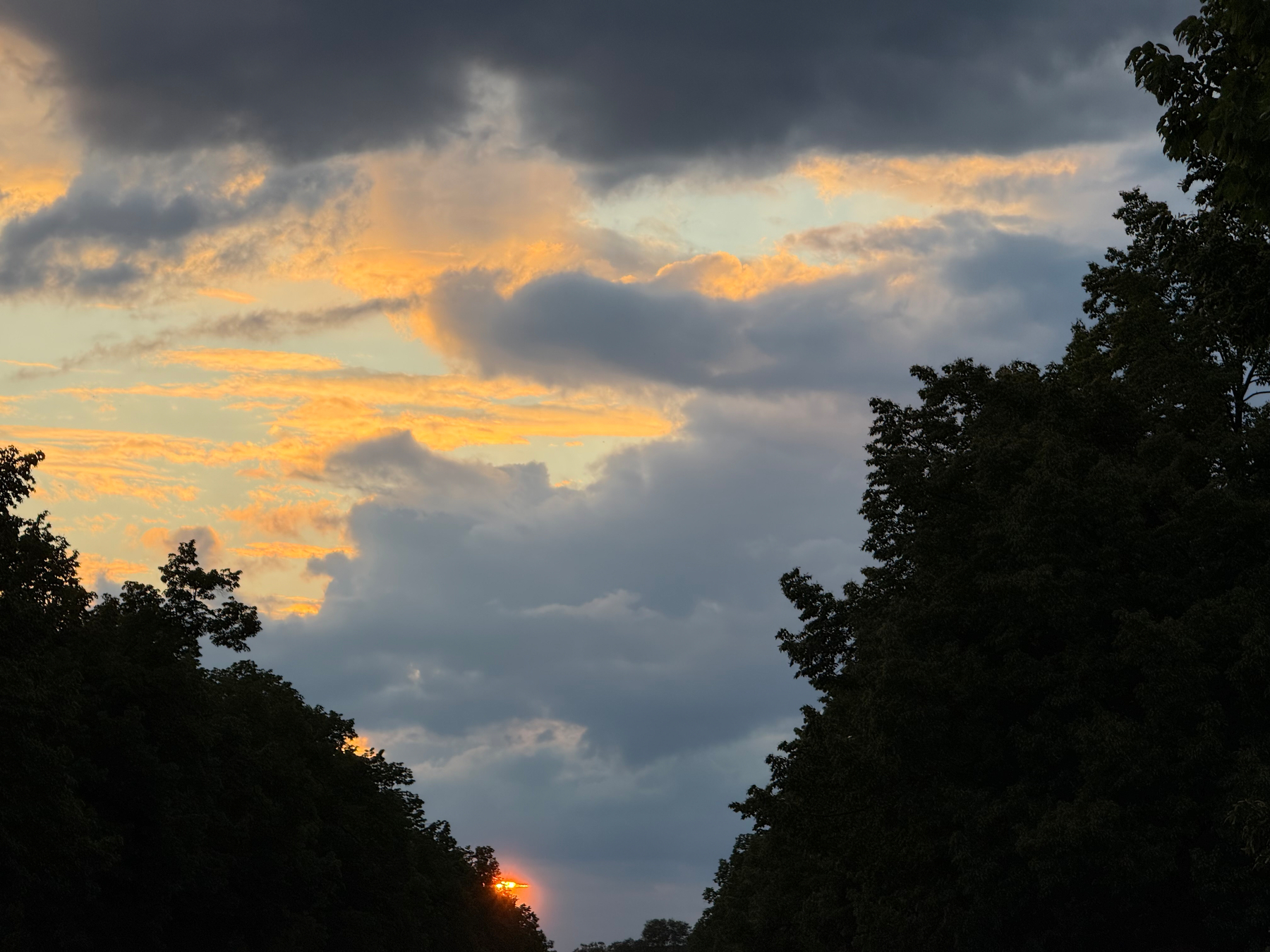 A dramatic sky with dark clouds partially shrouds a vivid sunset with a spot of bright orange sunlight, flanked by silhouettes of trees on either side.