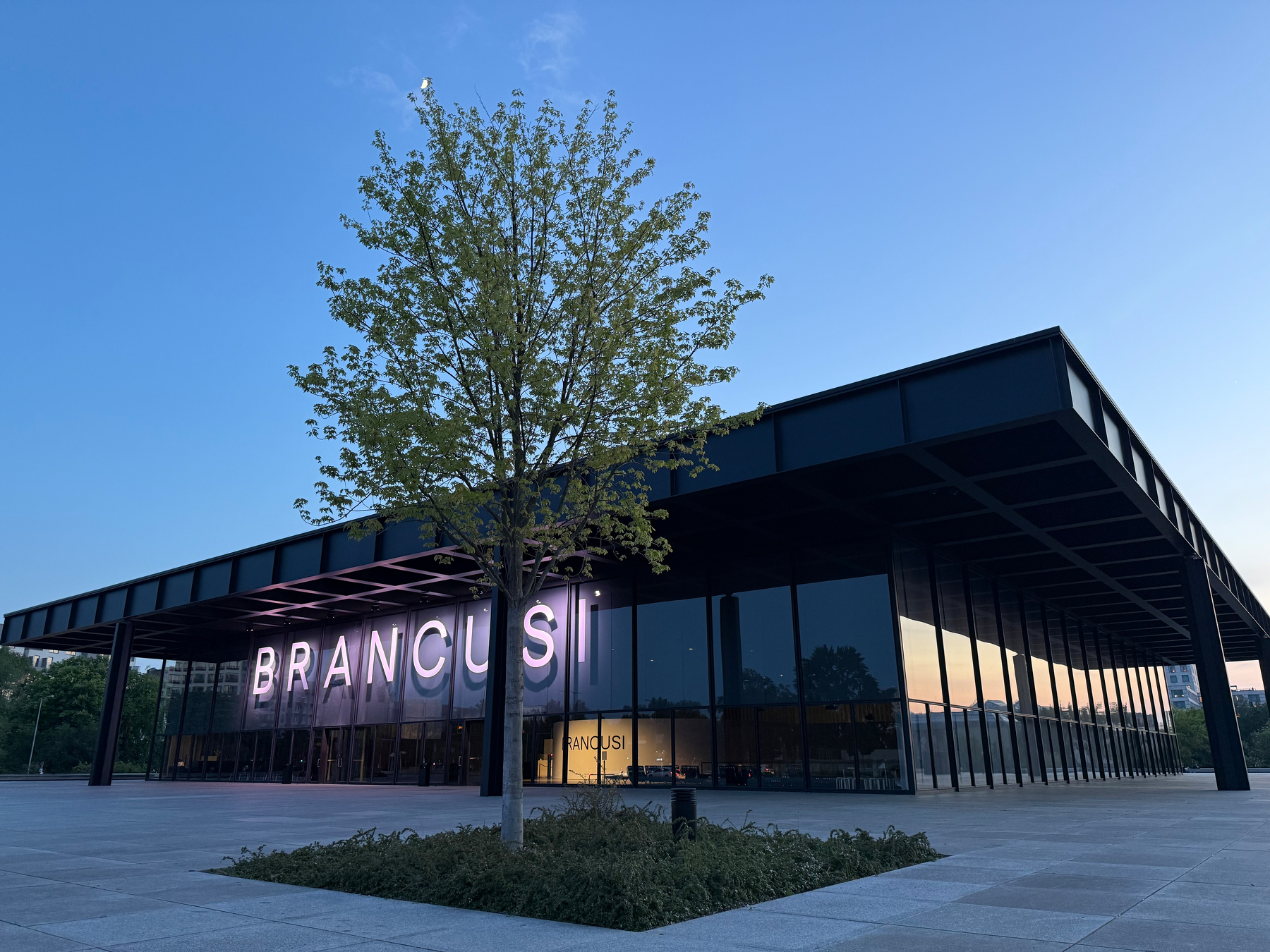 The modern glass and metal building of Mies van der Rohe’s Neue Nationalgalerie with a light sign displaying BRANCUSI is partially obscured by a tree in the foreground with quarter Moon and late sunset sky above.