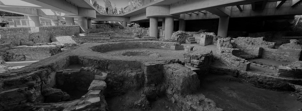 Ancient stone ruins with circular and rectangular structures are situated beneath modern concrete bridges.