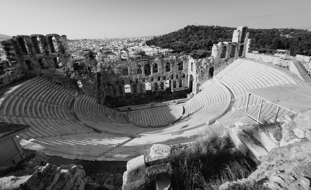 An ancient stone amphitheater with tiered seating overlooks a cityscape and hills in the background.