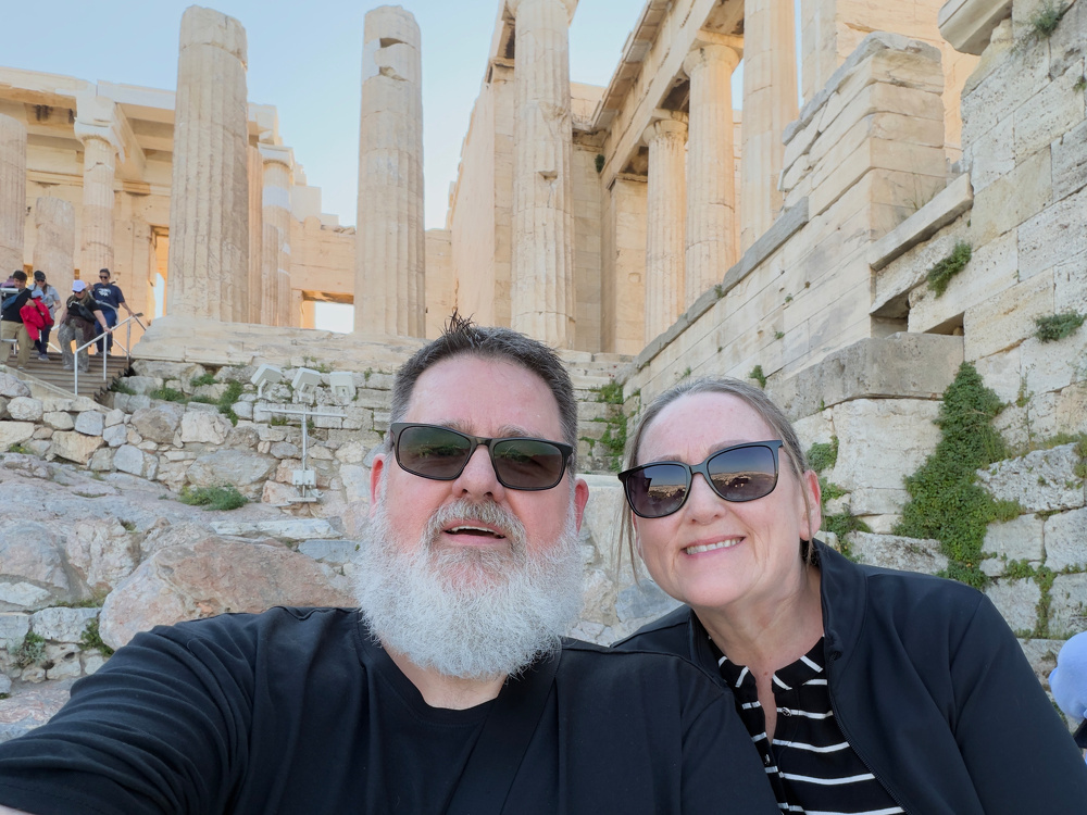 A man and a woman wearing sunglasses are taking a selfie in front of ancient ruins with large columns.
