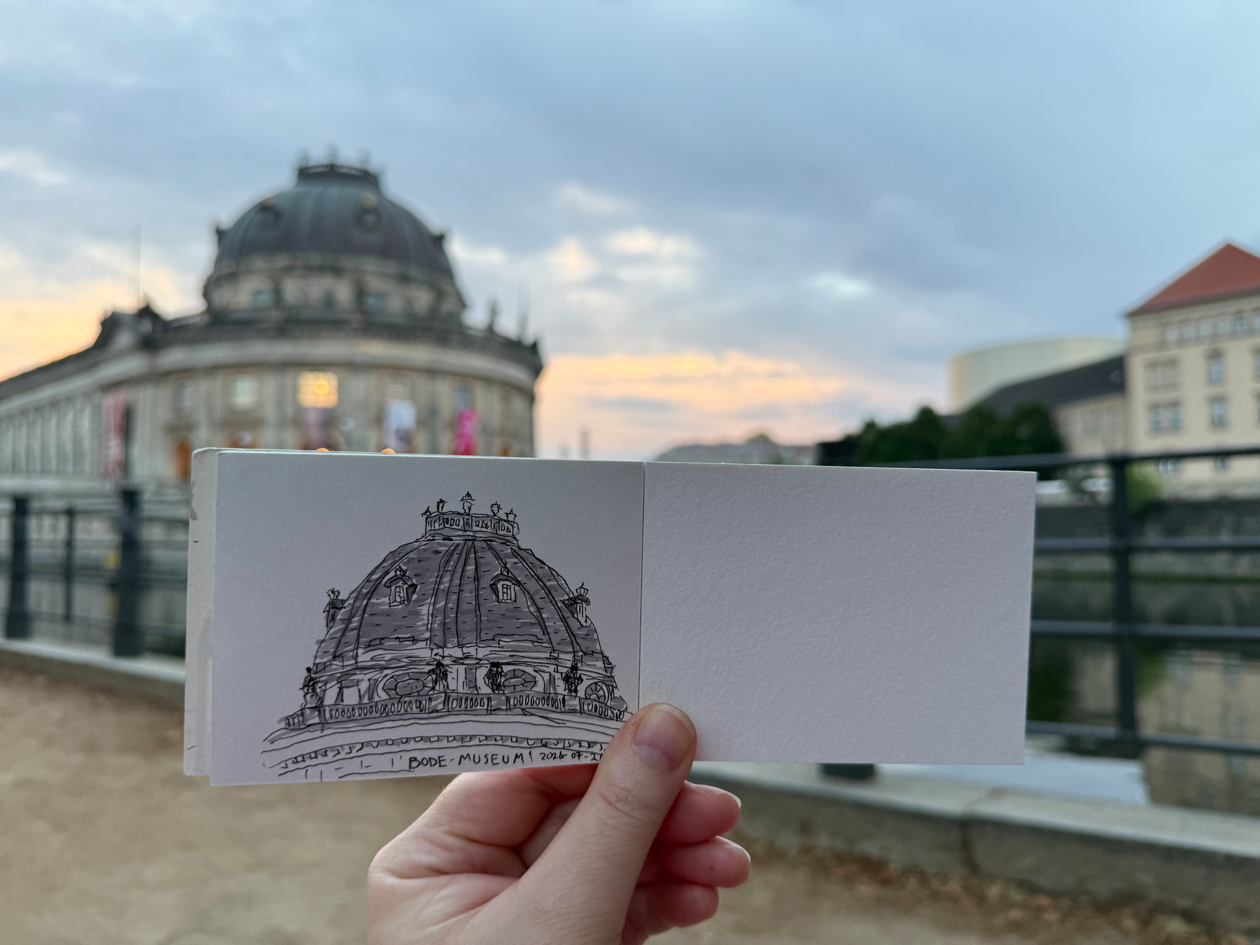 Eine Hand hält eine Zeichnung von einer Kuppel vor dem Gebäude mit Fluss und Sonnenuntergang.
