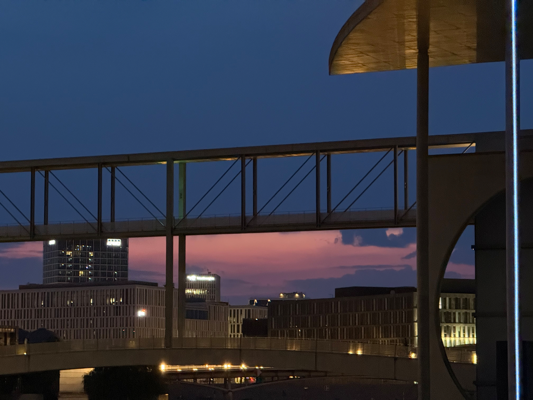 Eine Stadtlandschaft mit beleuchteten Regierungsgebäuden und einem Fußgängersteg ist bei Dämmerung unter einem blau-violetten Himmel zu sehen.