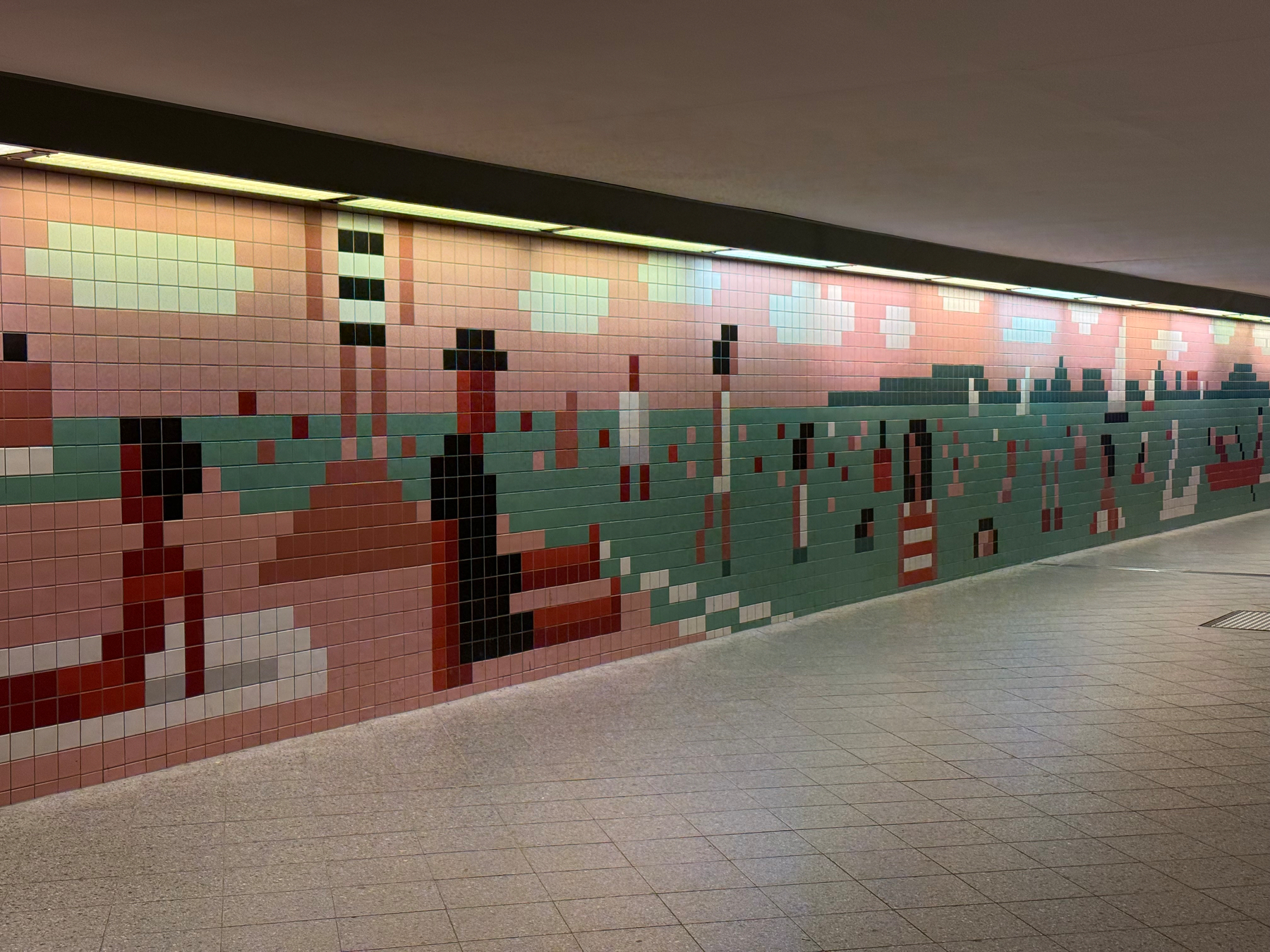 An einer langen Wand in einem Tunnel befindet sich ein Mosaik aus bunten Kacheln, das Menschen und einen Strand in stilisierter Form darstellt.