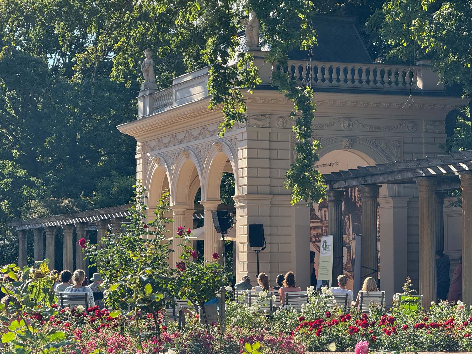 In einem sonnigen Park sitzen Menschen auf Bänken vor einem Pavillon mit Bögen und Skulpturen, umgeben von blühenden Rosen.