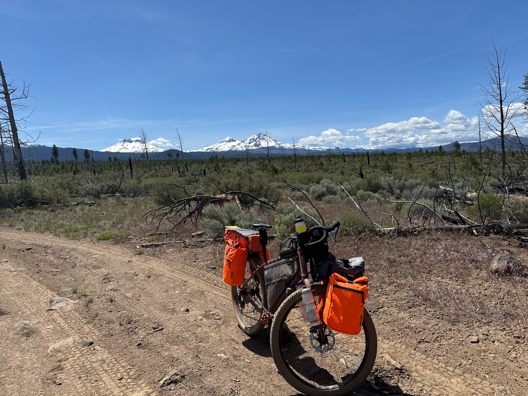 A bicycle with orange panniers is parked on a dirt path in a vast, barren landscape with snow-capped mountains in the distance.
