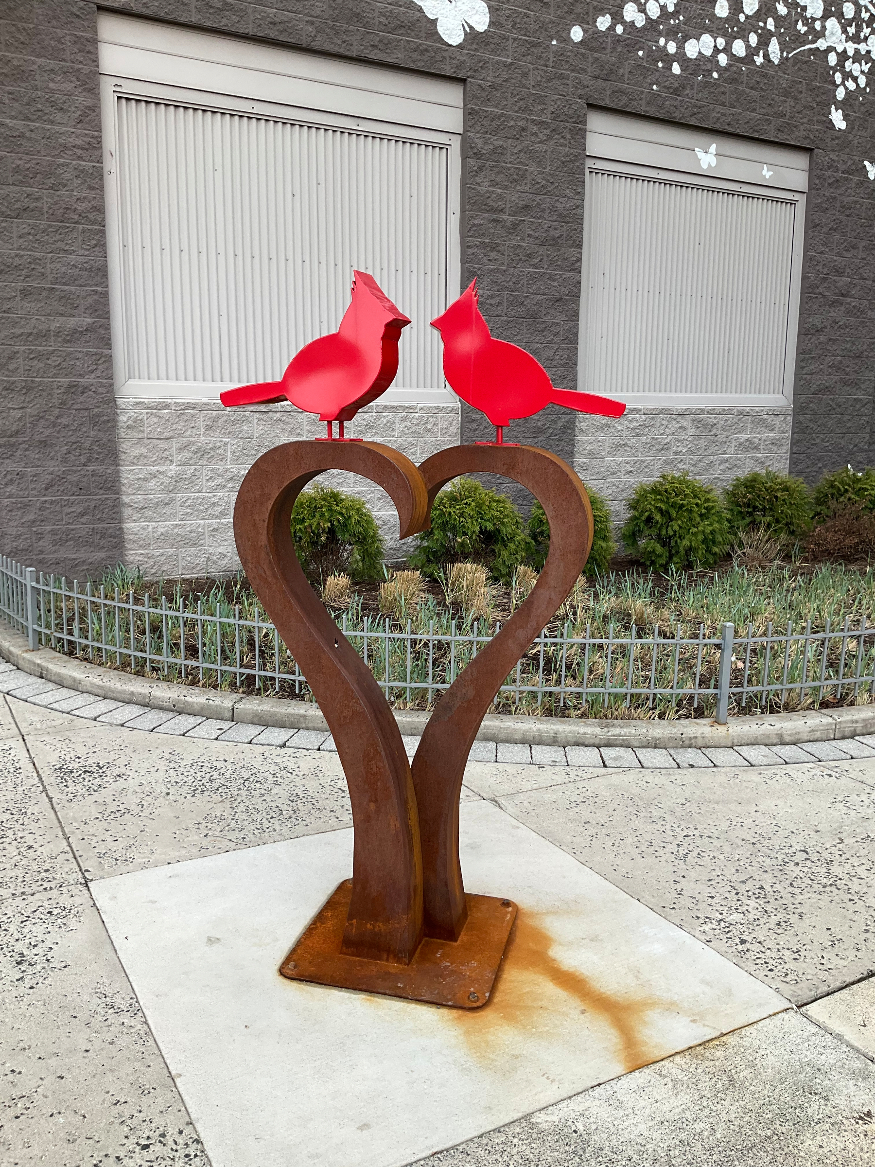 Steel sculpture with two curved branches coming up from the ground and forming a valentine shape. On top are two cardinals, beak to beak. Both cardinals are bright red. 