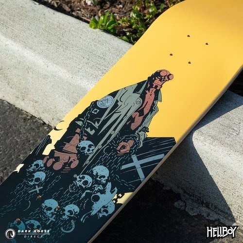 Auto-generated description: A skateboard deck features artwork of Hellboy standing among skulls and chains against a yellow background.
