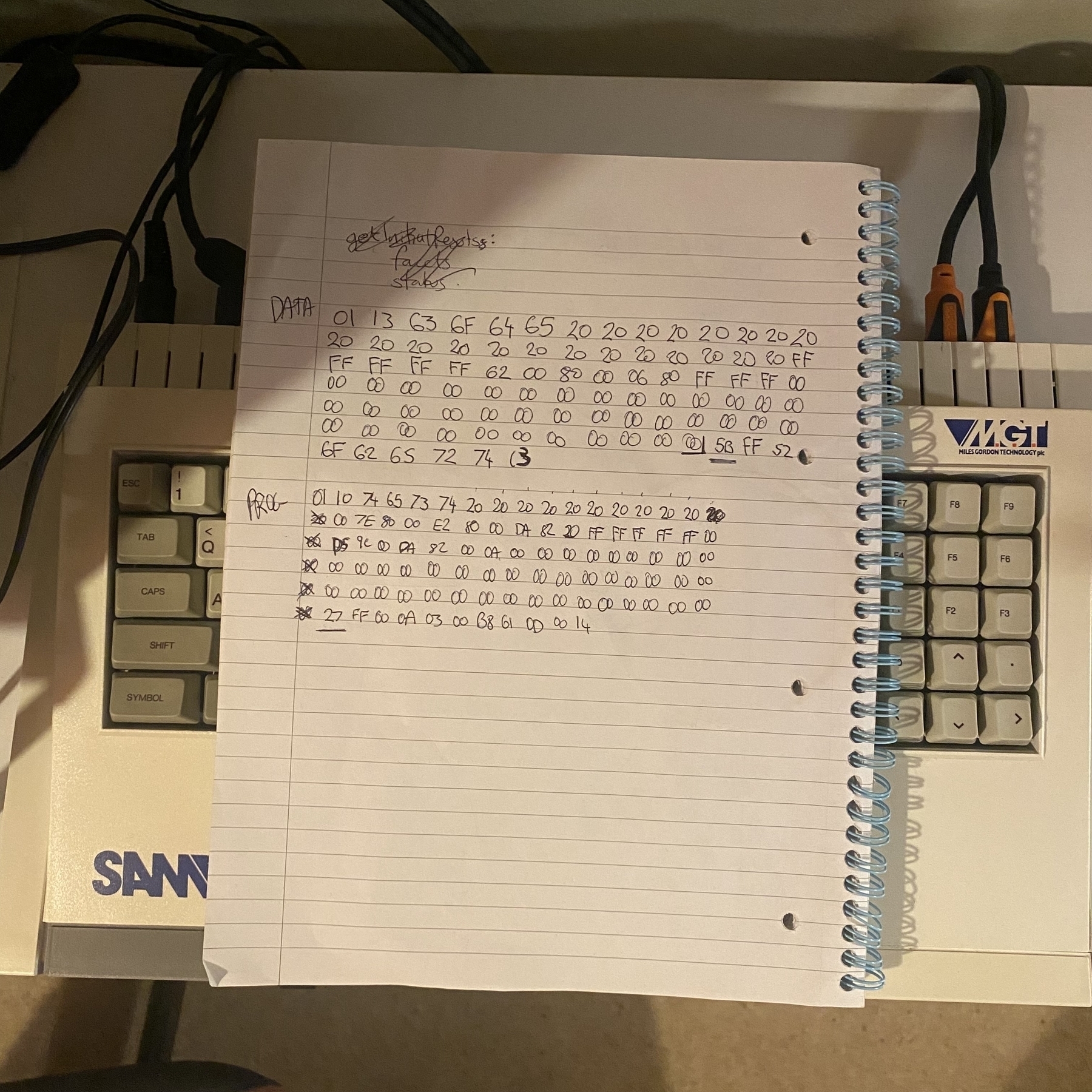 Handwritten hexadecimal numbers on a lined notepad. The notepad is on top of SAM Coupé