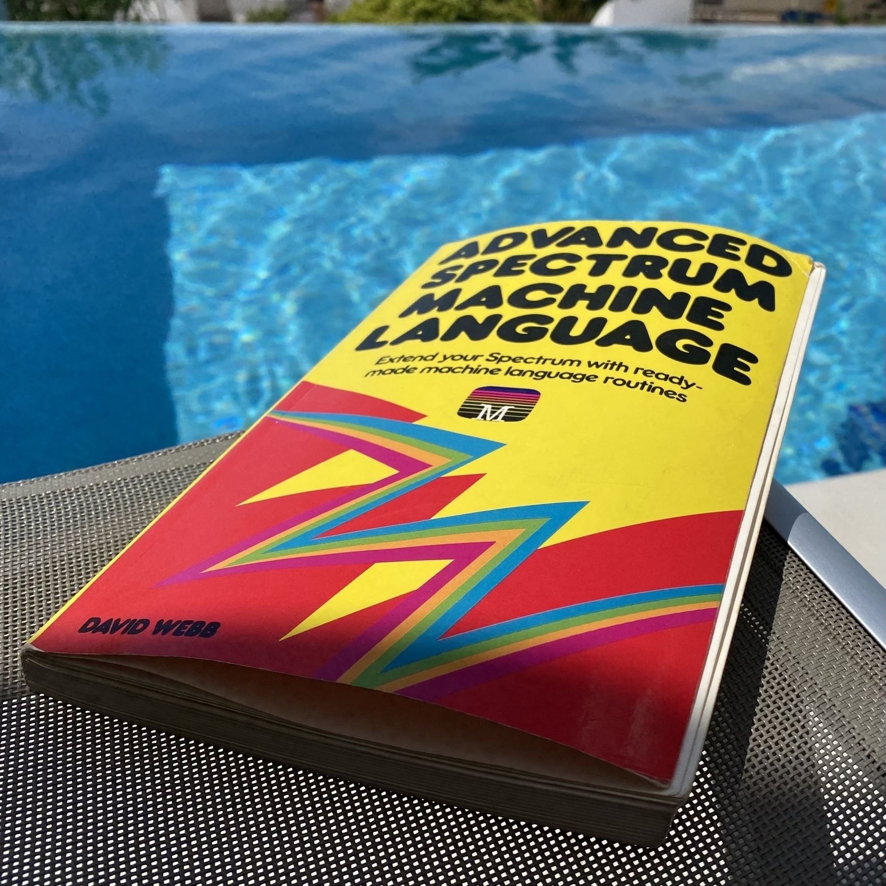 Paperback book in the sunshine by a swimming pool. The title is Advanced Spectrum Machine Language, by David Web.