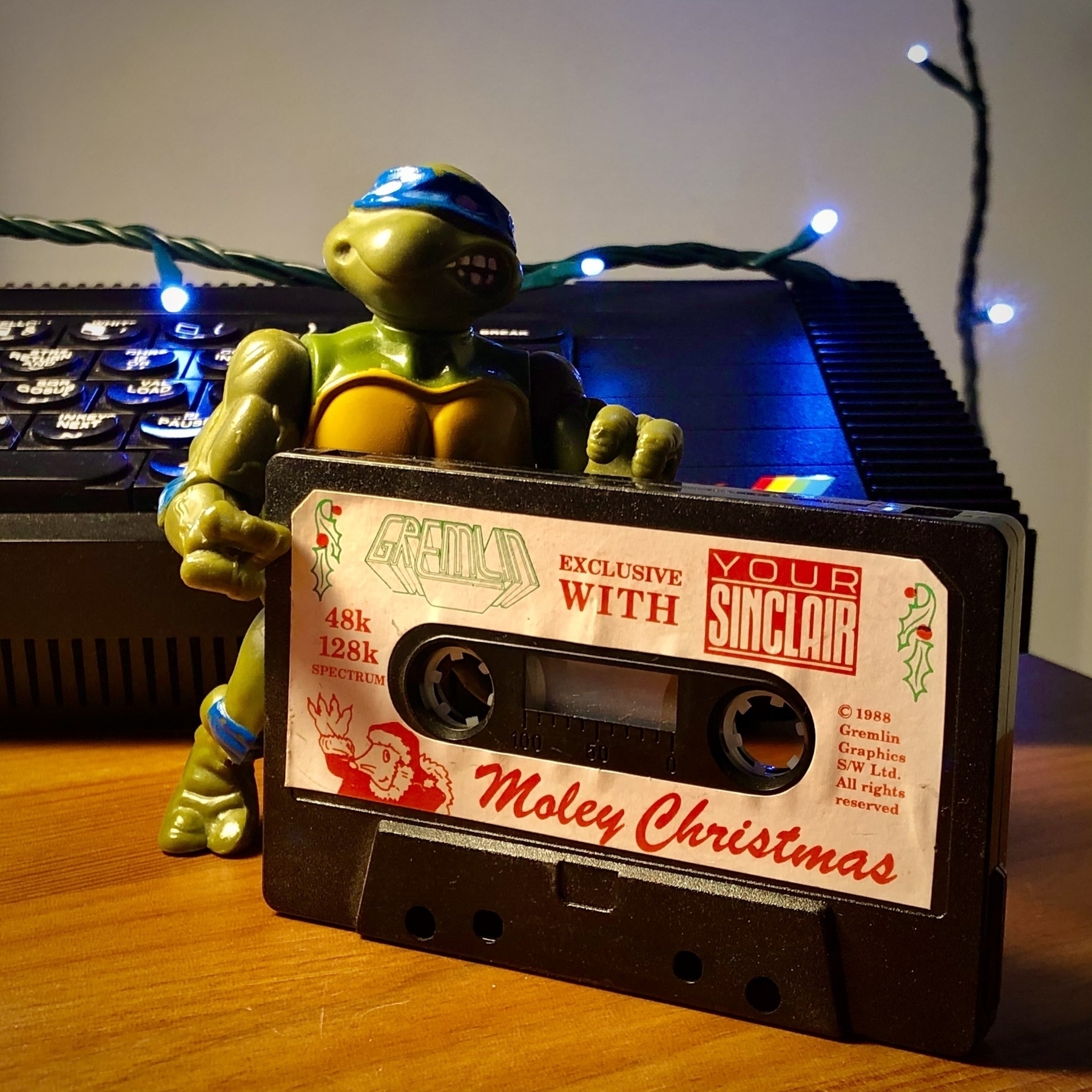 “Moley Christmas” game for ZX Spectrum on cassette tape being held by Leonardo (Teenage Mutant Ninja Turtles) action figure, standing in front of a ZX Spectrum. Fairy lights in the background.