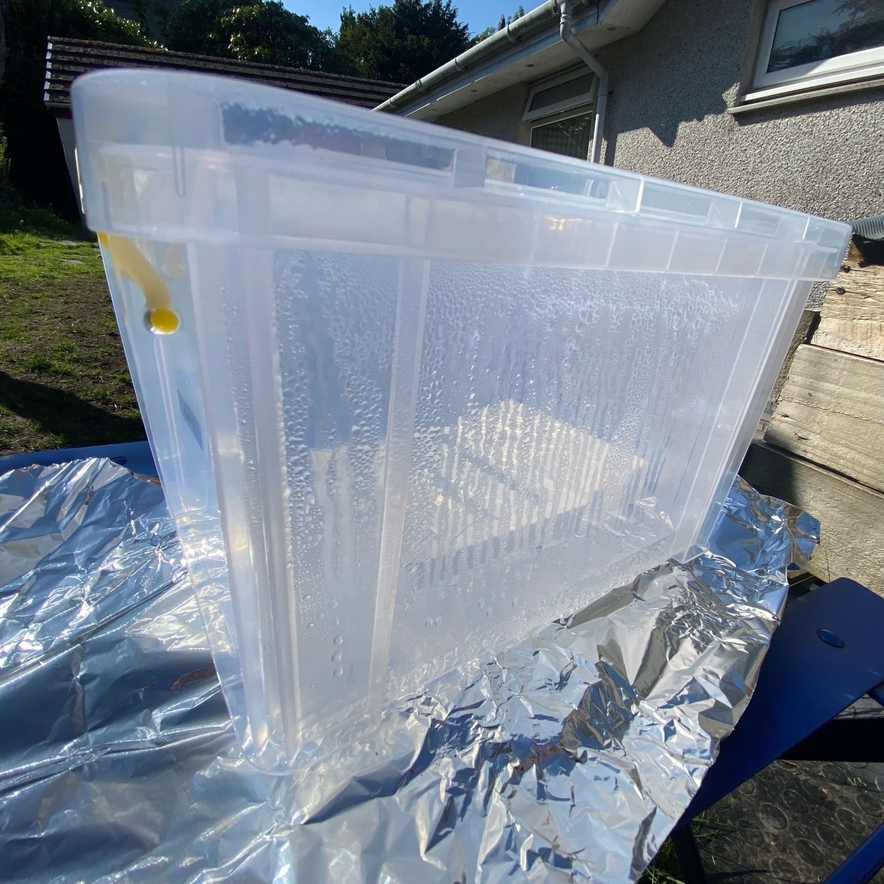 a clear Perspex™ box in direct sunlight with a white plastic item inside. aluminium foil underneath, the clear liquid in the box is forming condensation on the walls and lid