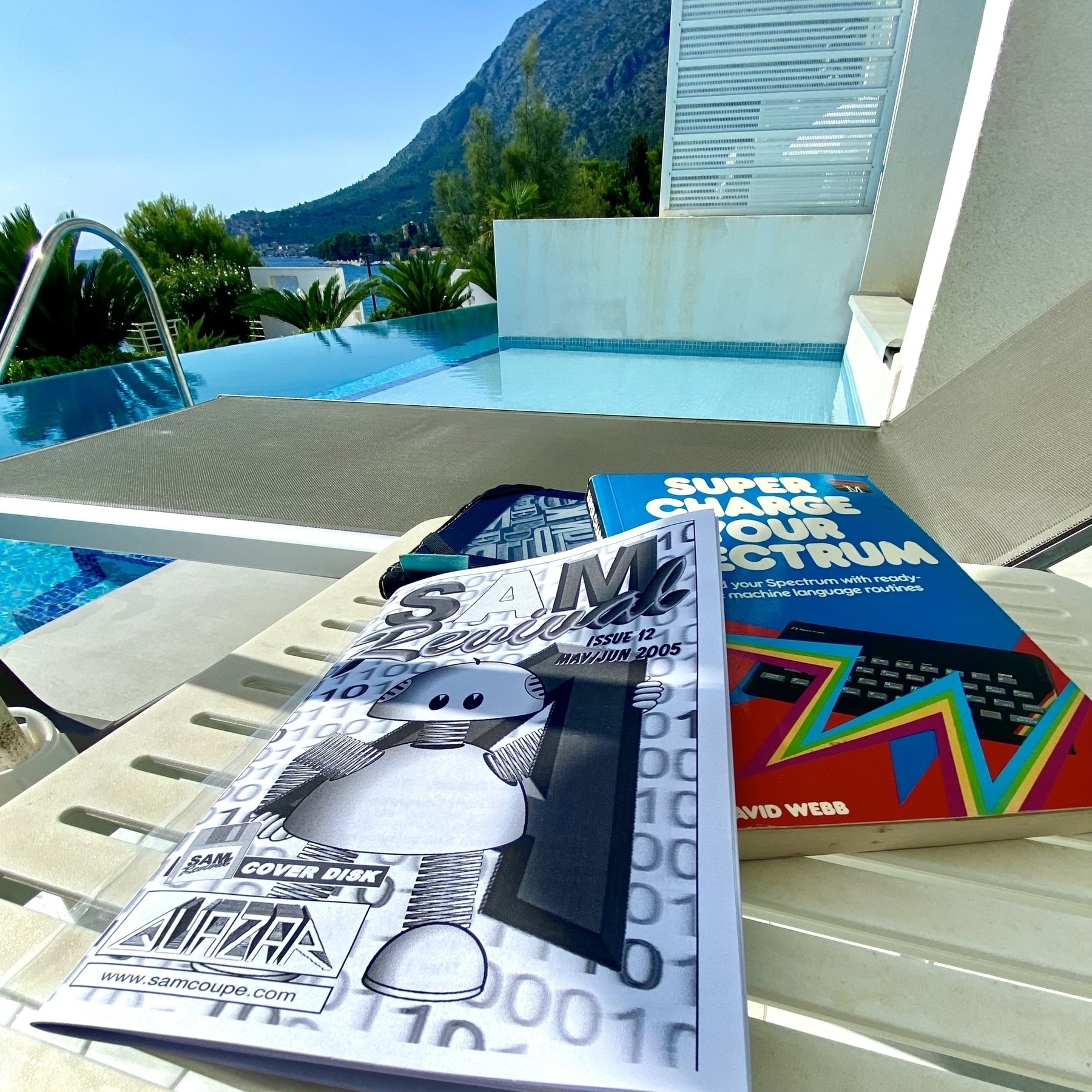 Literature by a swimming pool. “SAM Revival” magazine by Quazar, and “Super Charge Your Spectrum” by David Webb. Palm trees and steep mountains are in the background.