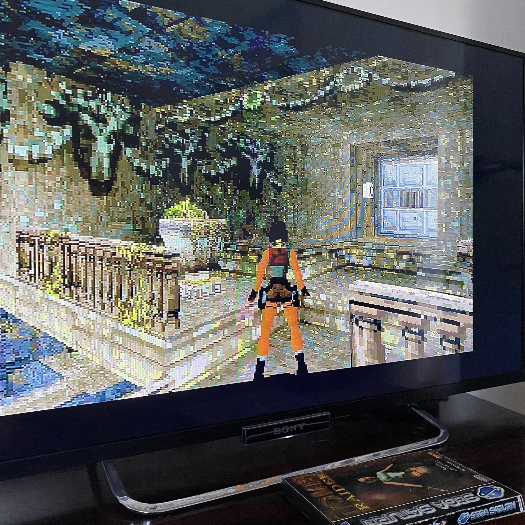 Screenshot of NTSC version of Tomb Raider, the Cistern level. Lara Croft is standing on a stone balcony looking at a disguised moveable block that’s hiding a secret area. Unlike the PAL version the texture of the block is the same as the surrounding walls so it’s harder to find. (Note the screen borders as this is running on my 50Hz PAL Saturn!)