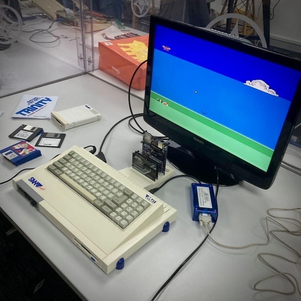 SAM Coupé computer with a few expansion cards plugged in, connected to an LCD television. On the screen is a blue and green horizon. Disks (and a spare SAM Drive) are scattered around.