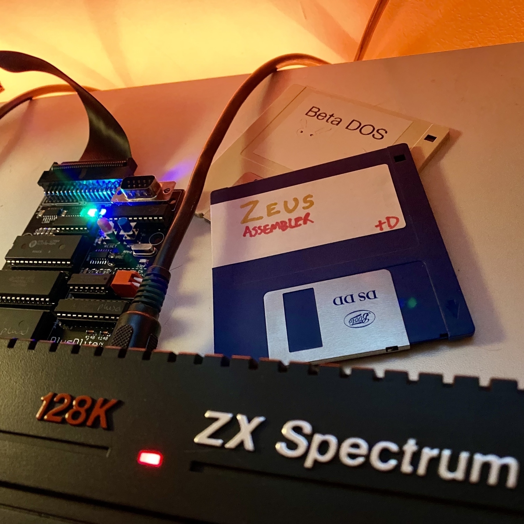 PlusDLite disk interface without its casing, plugged into a ZX Spectrum +2A. A couple of disks are scattered around with labels “Zeus Assembler” and Beta DOS”.