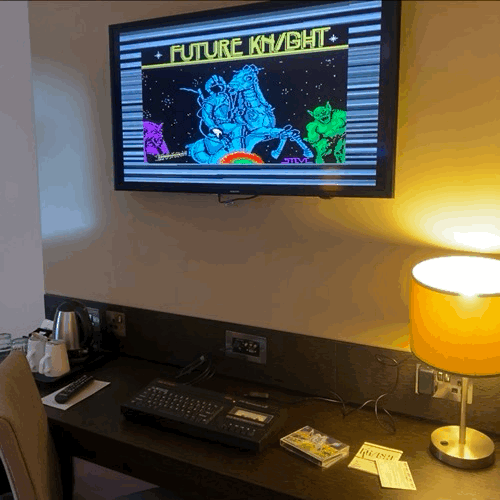 Animated GIF. Image shows a wall mounted hotel room TV displaying the loading screen of Future Knight on ZX Spectrum (the computer is on the desk loading the game from cassette tape).