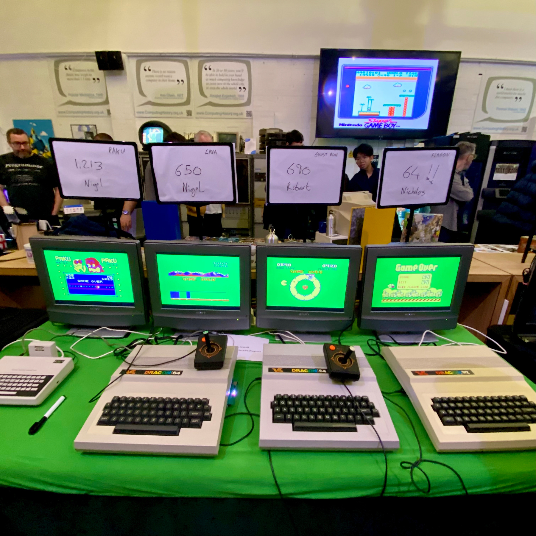 Photograph of micro computers on a table.  Three dragons (and a CoCO?) Each computer has a different game, above each display is a high score dry-erase board with scores by Nigel, Robert and Nicholas. 