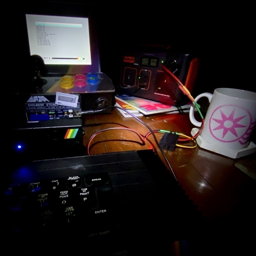 Photograph showing a computer, screen, battery pack and a mug illuminated by torchlight and the glow of the small screen, in the dark. 