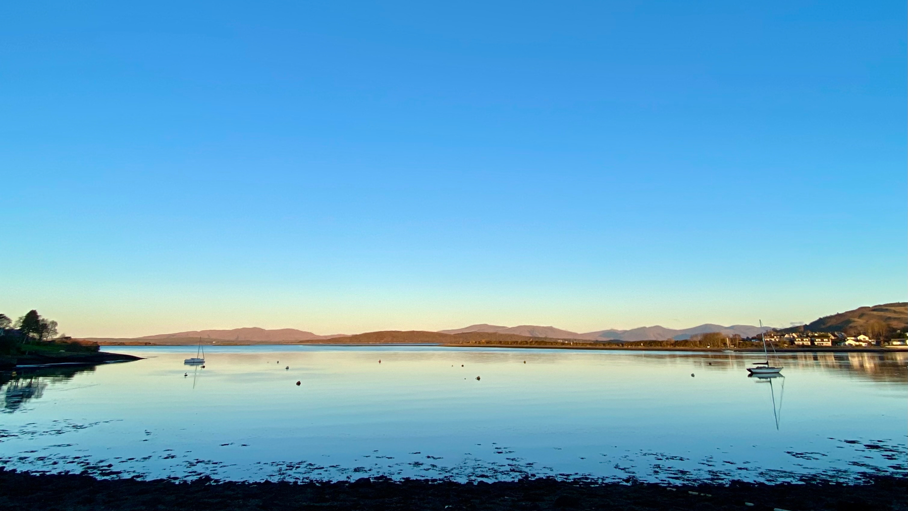 Photograph of Loch Etive on a clear November morning. The sky is blue and the water is still. A couple of moored sailing boats are visible; as is North Connel on the opposite shore.