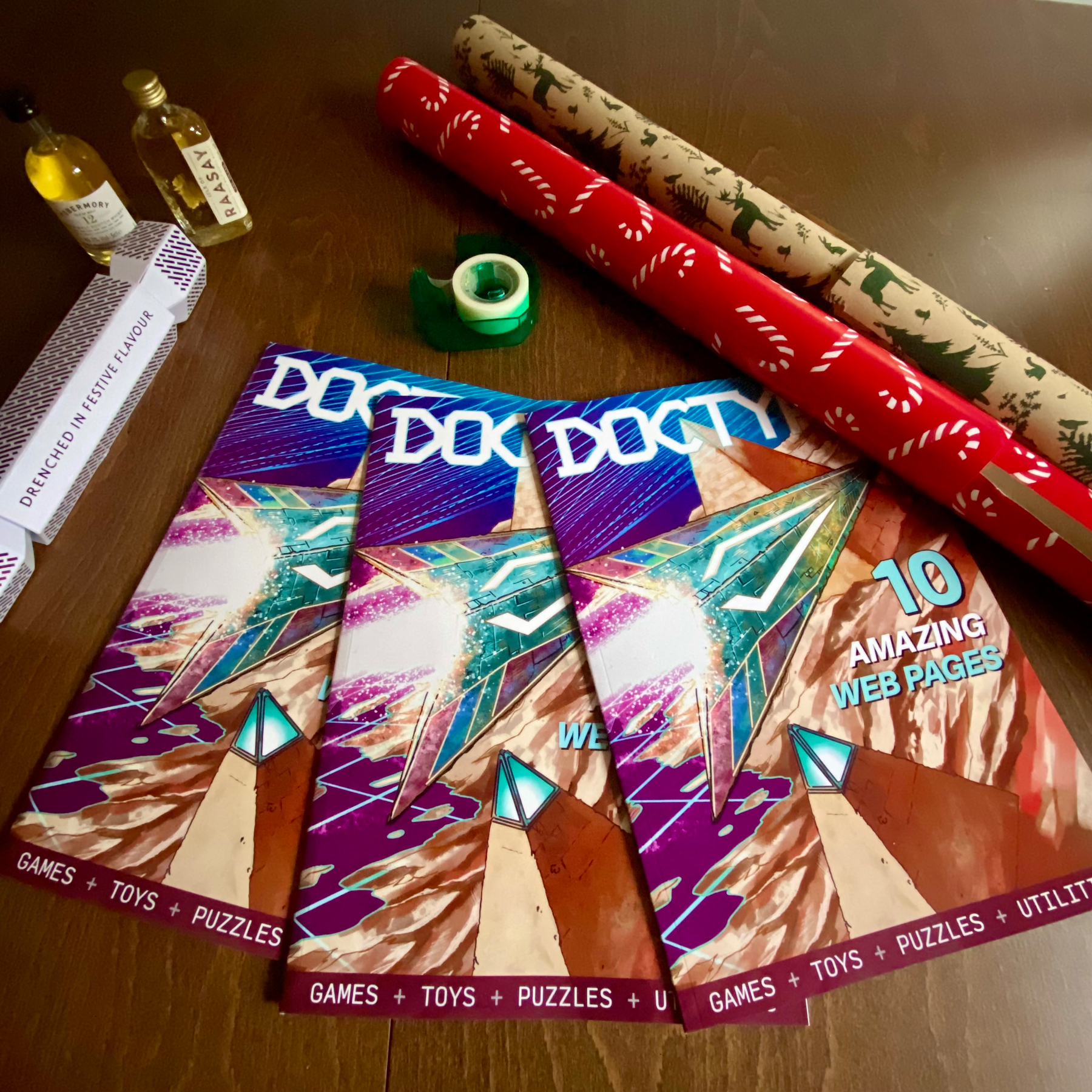 Photograph of three copies of DOCTYPE magazine on a tabletop with Christmas wrapping paper and some unidirectional bondage tape. Also some Scottish islandy booze. 