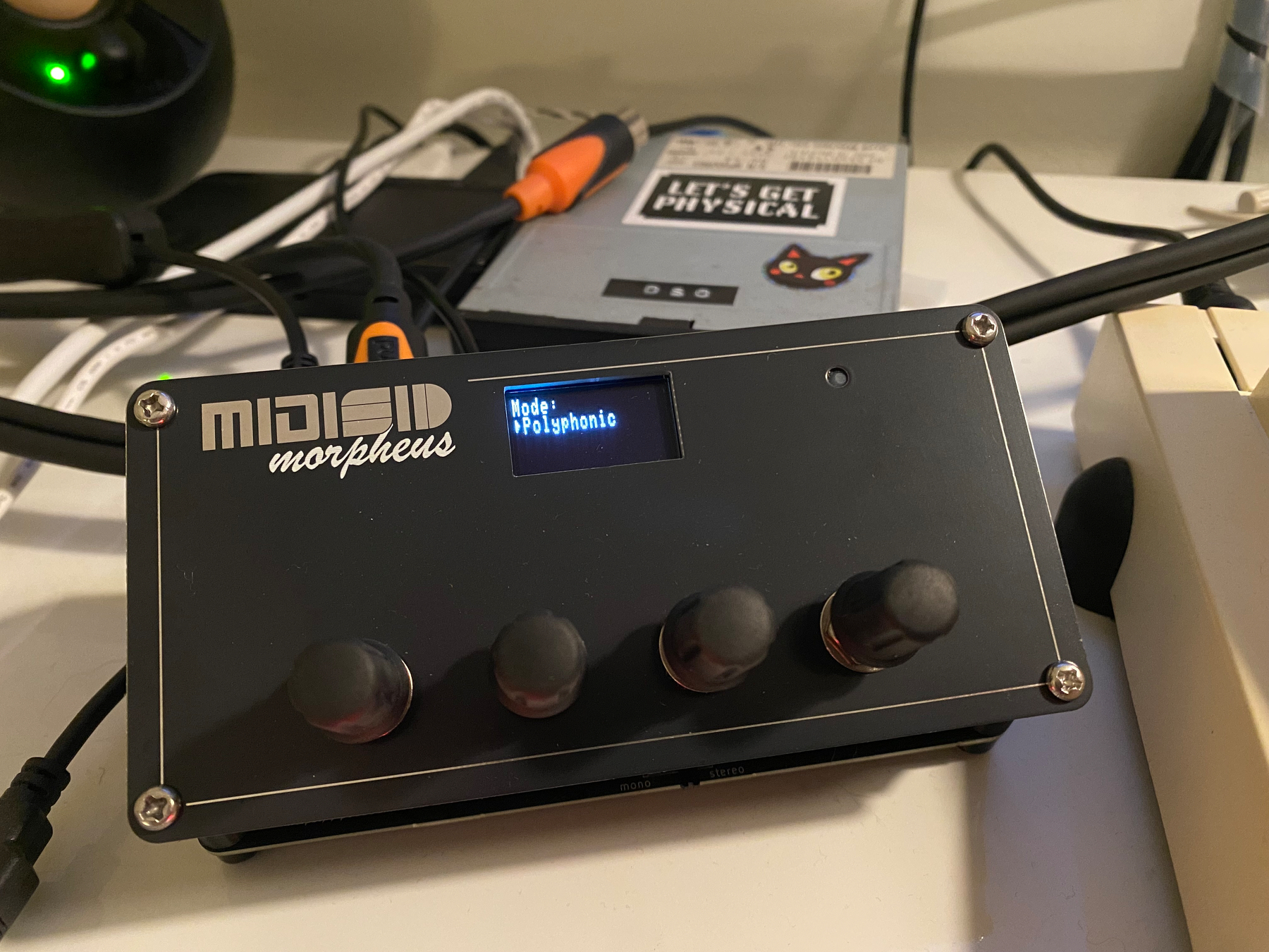 Photograph of MIDISID Morpheus, a neat oblong black box with a small screen and four twiddly knobs. The device is attached to various wires.