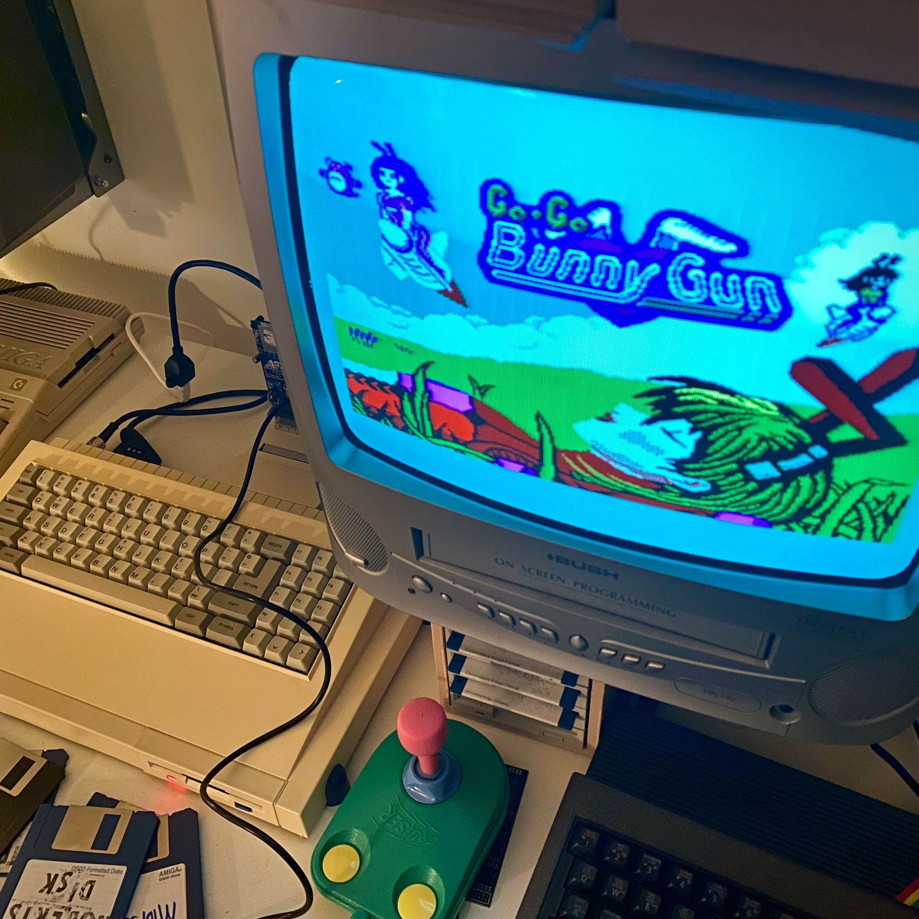 A photograph of the BunnyGun splash screen displayed on a CRT telly running on SAM Coupé. Also pictured is a lurid green, pink and yellow joystick and several floppy disks.