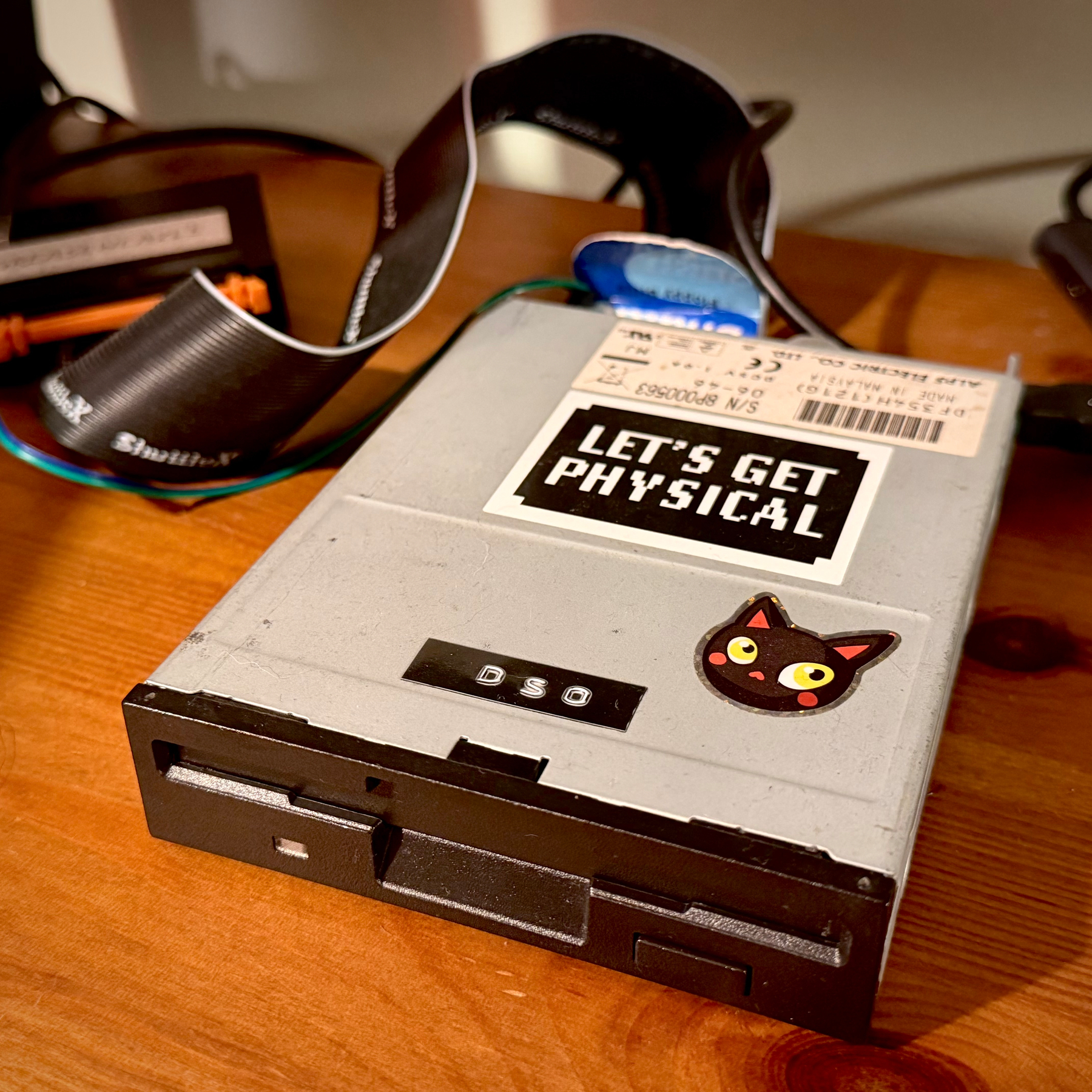 Photo of a pretty standard PC 3.5" diskette drive connected via ribbon cable to a Greaseweazle doodah. Someone really cool has put stickers on the disk to prove how cool they are. Text on the stickers reads “LETS GET PHYSICAL” and “DS0” (hinting that the drive is wired to the Shugart standard rather than the more common “IBM PC” way of doing things. Did I mention the owner of this gear is really cool?)