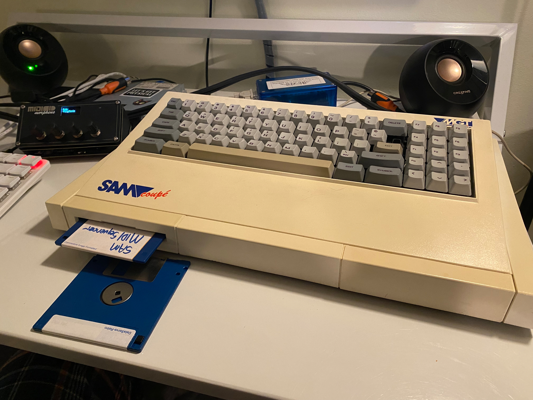 Photograph of SAM Coupé with a floppy disk labelled “SAM MIDI Sequencer”. The MIDISID and some small speakers are also on the same desk. 