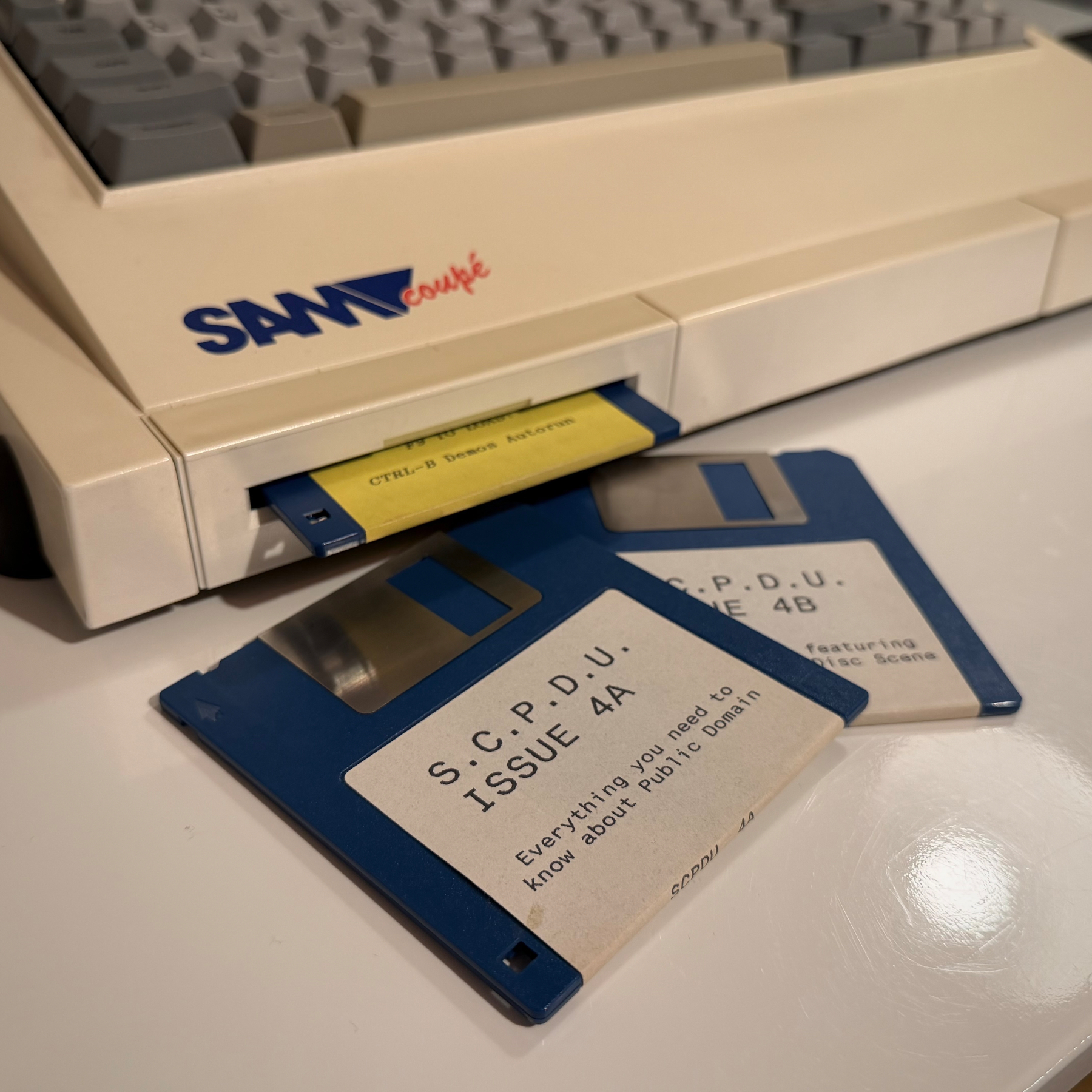 Photograph of SAM computer with a disk partially inserted into the floppy disk drive, and two more disks on the desk in front of the machine. Very carefully staged and photographed to give the impression of ineffably cool nonchalance. Text on the frontmost disk reads “S. C. P. D. U. ISSUE 4A, Everything you need to know about Public Domain”