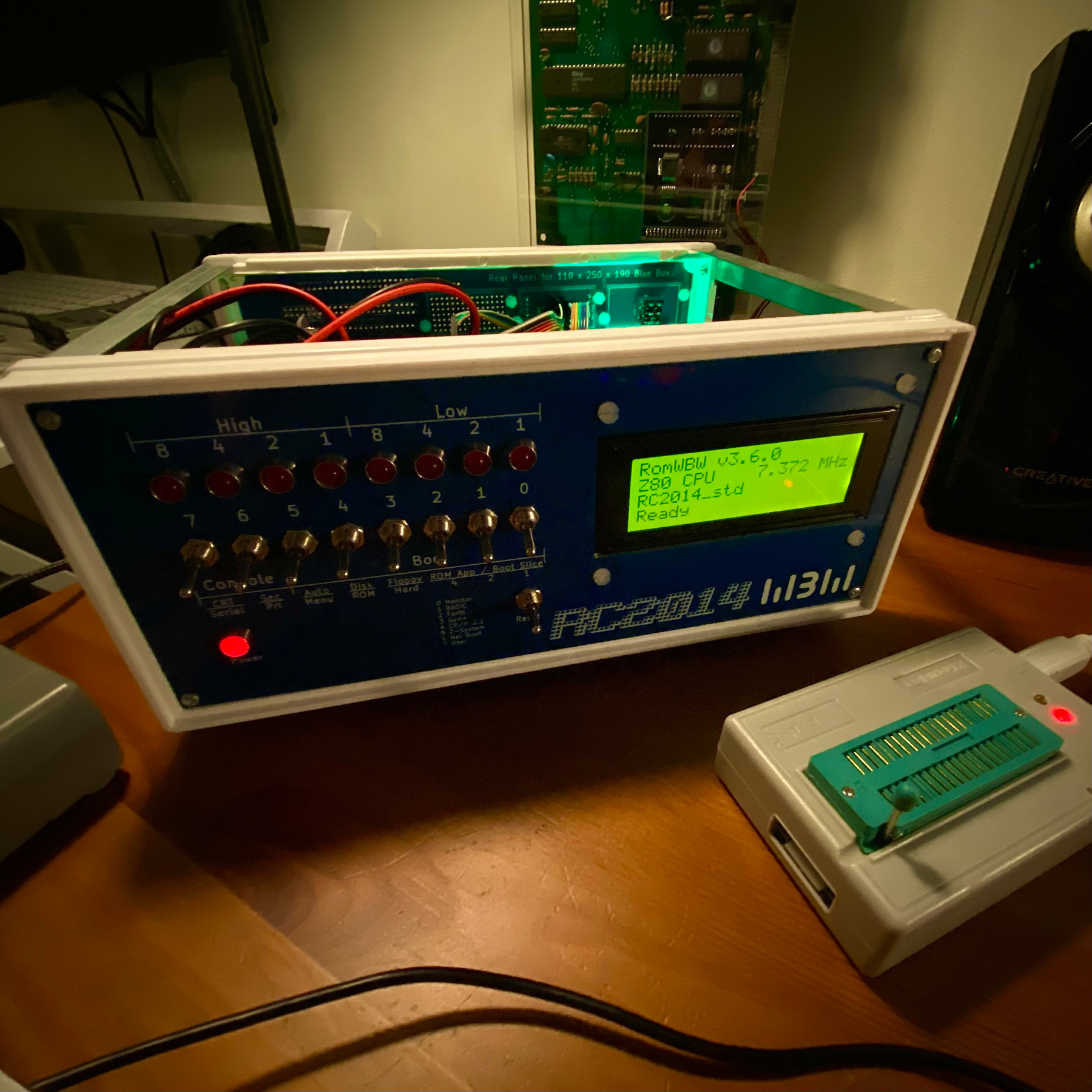 Photograph of a little blue box that appears to be bigger on the inside. The front panel is adorned with toggle switches, LEDs and a green LCD glowing brightly. Text on the small screen reads RomWBW V3.6.0, Z80 CPU 7.372 MHz, RC2014_std, Ready. Also pictured is a ROM programmer.