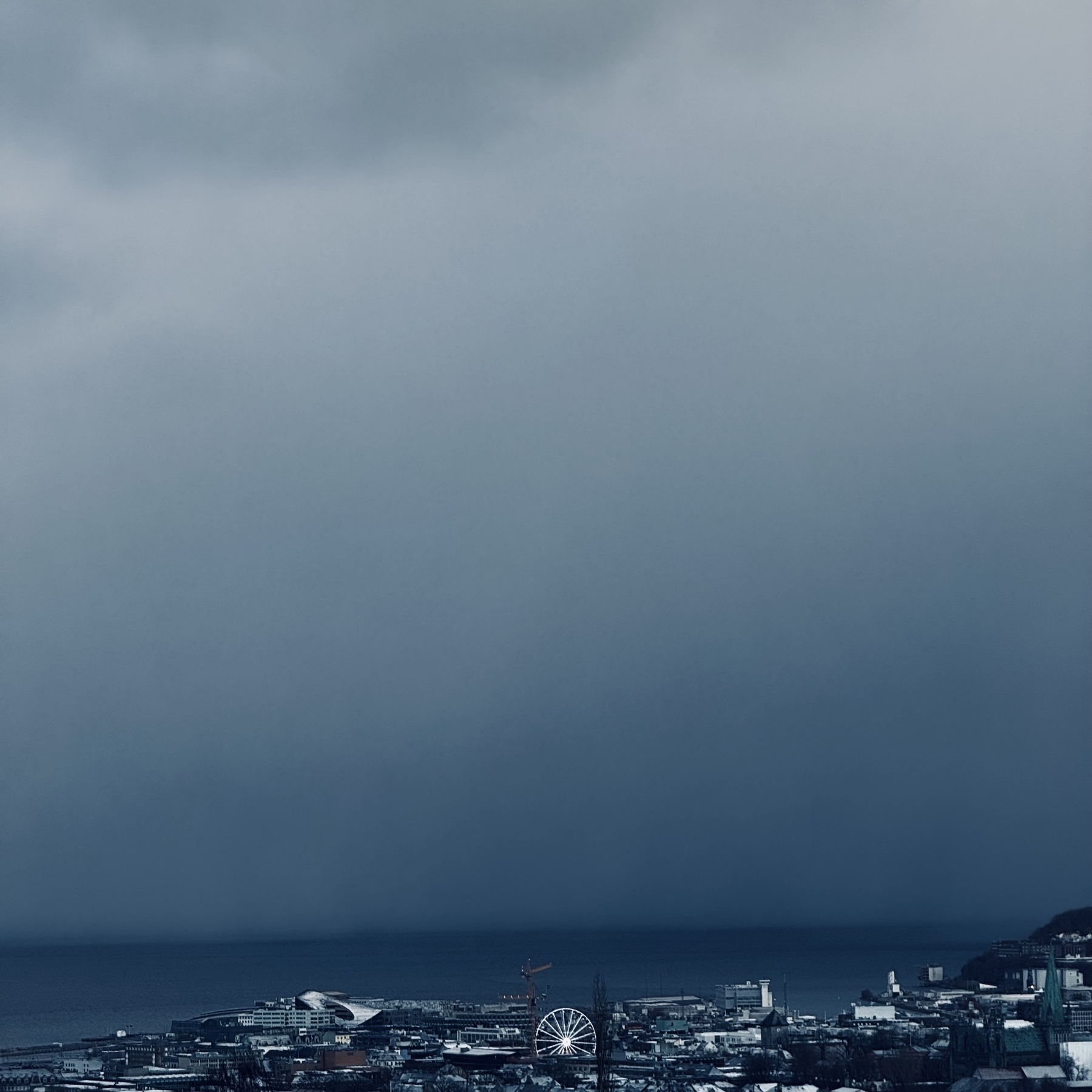 A cityscape features a large Ferris wheel under a dramatic, cloudy sky near a body of water.