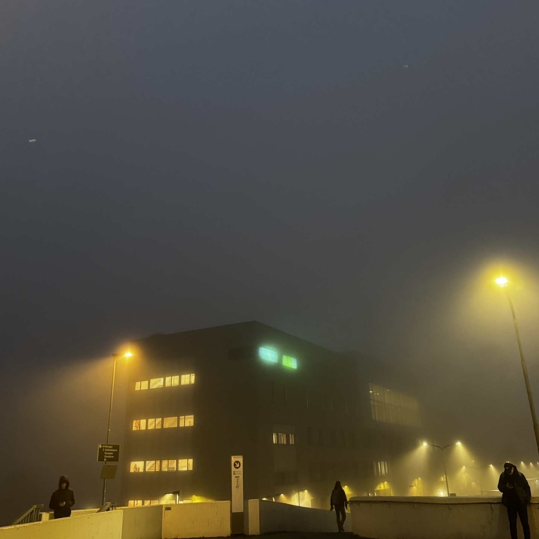 A foggy evening scene shows a street with glowing streetlights and a modern, illuminated building.