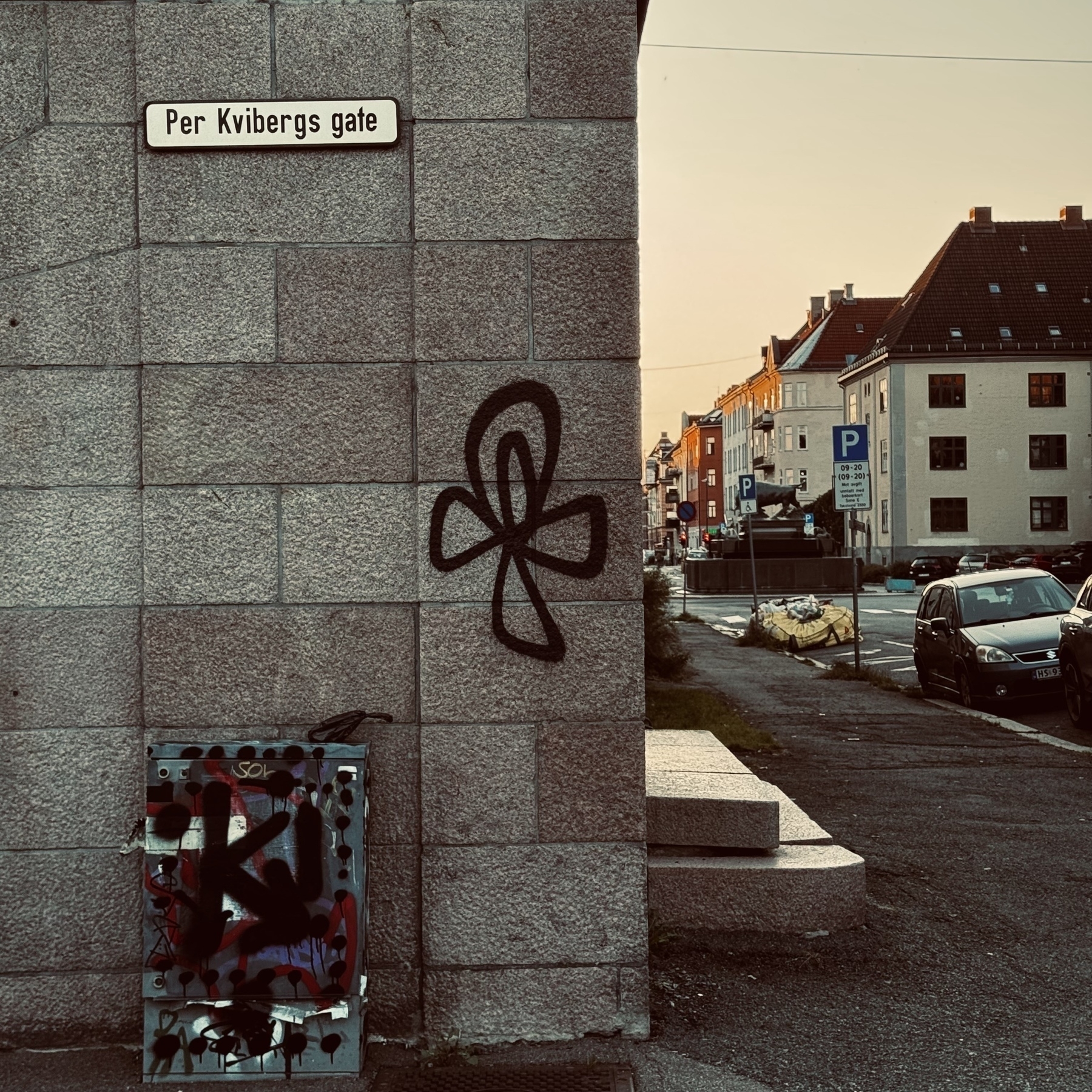 A street sign for "Per Kvibergs gate" is affixed to a building wall with graffiti, located alongside a road with parked cars and buildings.