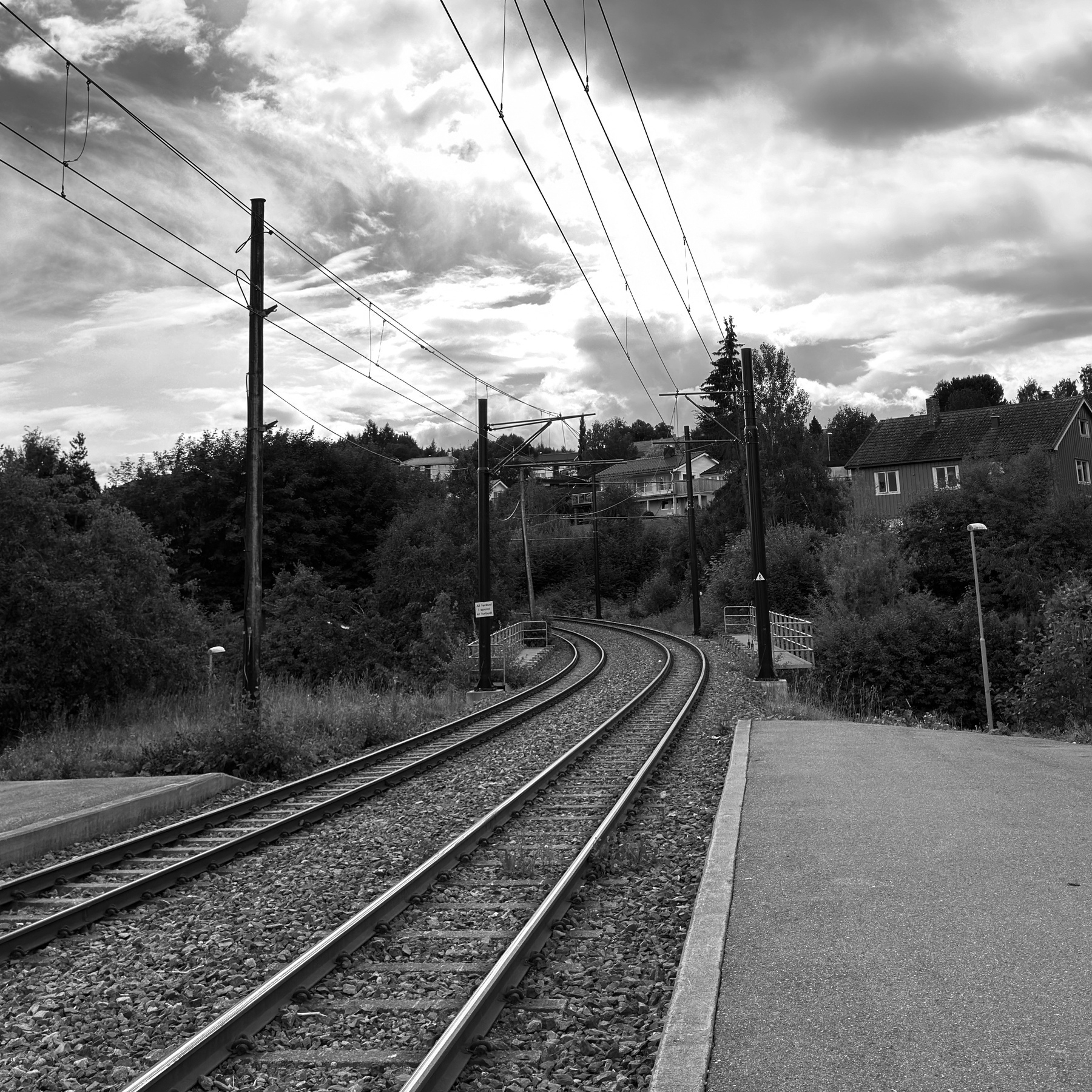 A tram track curves through a rural area with overhead power lines and surrounding vegetation under a cloudy sky.