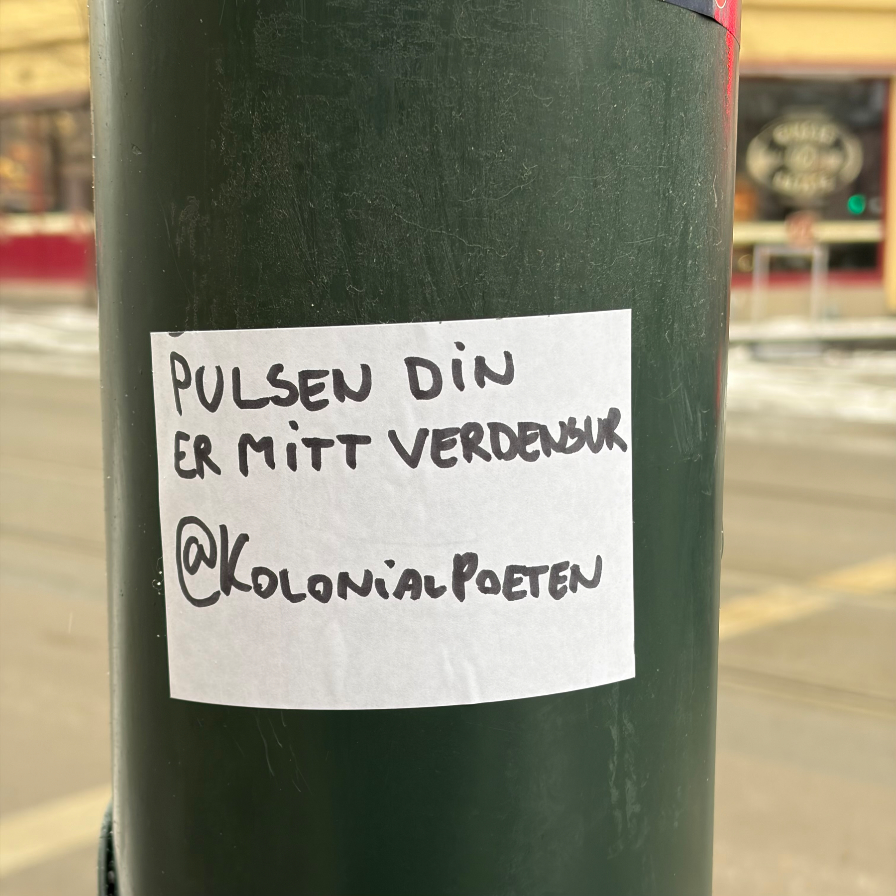En lapp festet til en stolpe med teksten PULSEN DIN ER MITT VERDENSUR @Kolonialpoeten.