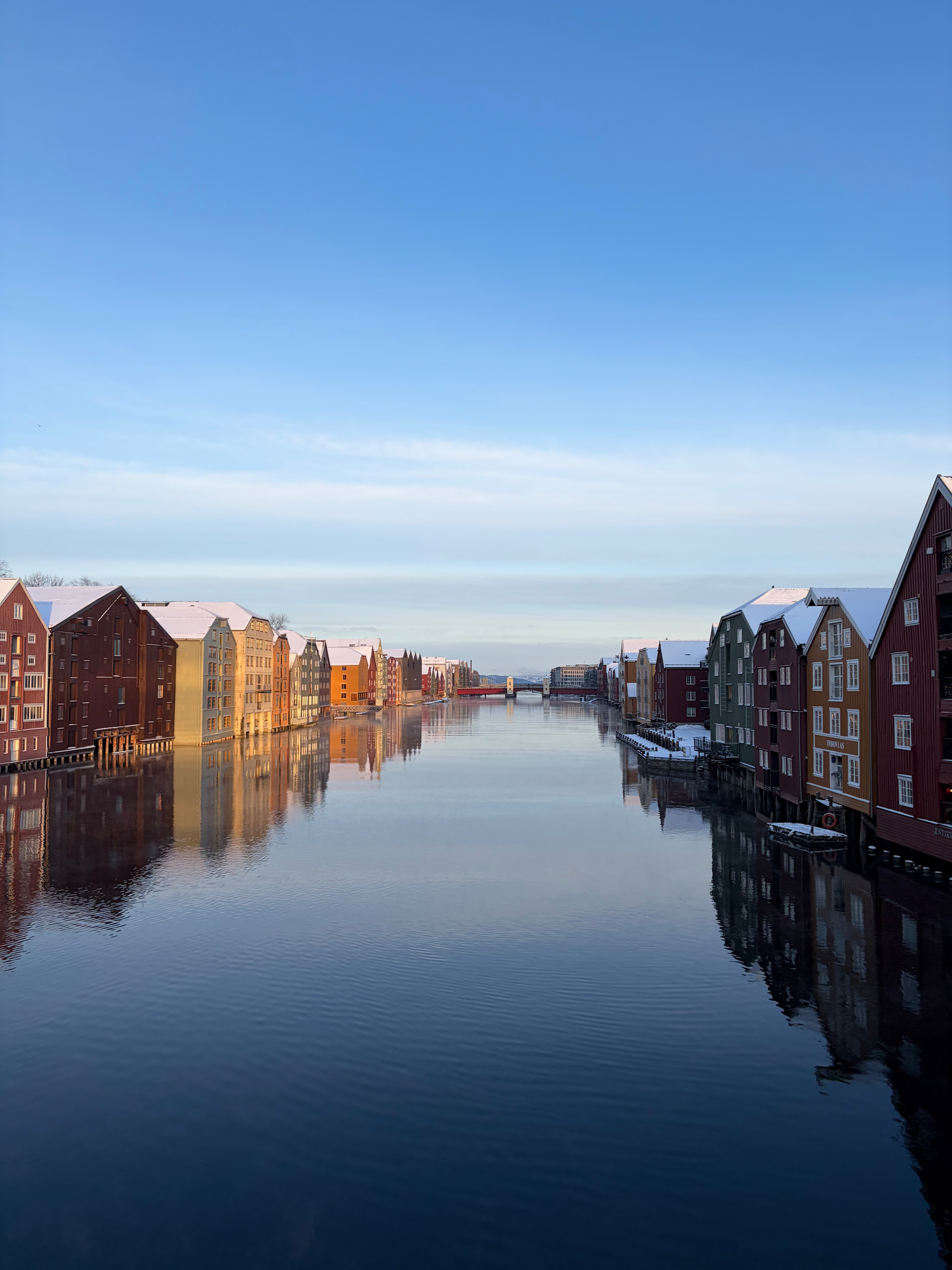 Tradisjonelle brygger i ulike farger speiler seg i en rolig elv under en klar, blå himmel.